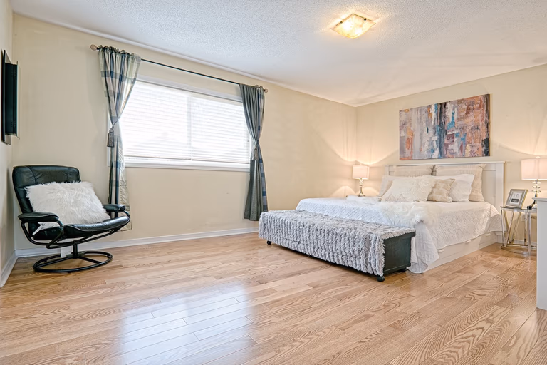 This spacious master bedroom features a simple and lovely interior design with natural hardwood flooring, soft neutral wall tones, and a well-balanced furniture layout. Symmetrical bedside lighting and minimal decor create a clean, relaxing atmosphere, while large windows provide ample natural light. The design focuses on comfort, openness, and timeless style, making it suitable for family homes and long-term residential living.