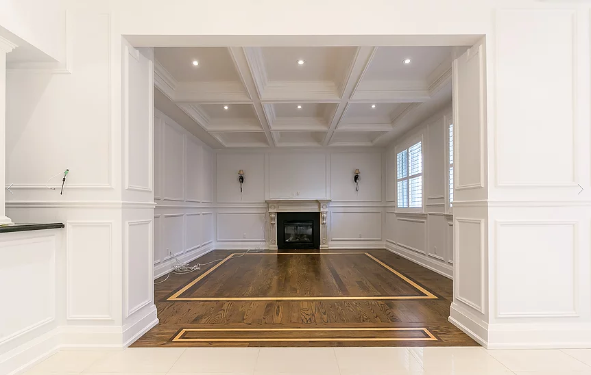 This Richmond Hill interior renovation highlights a refined living space with a coffered ceiling, custom wainscoting, and detailed hardwood flooring with inlay borders. Large windows with plantation shutters enhance natural light, while classic millwork and clean architectural lines create a timeless, elegant interior renovation result.