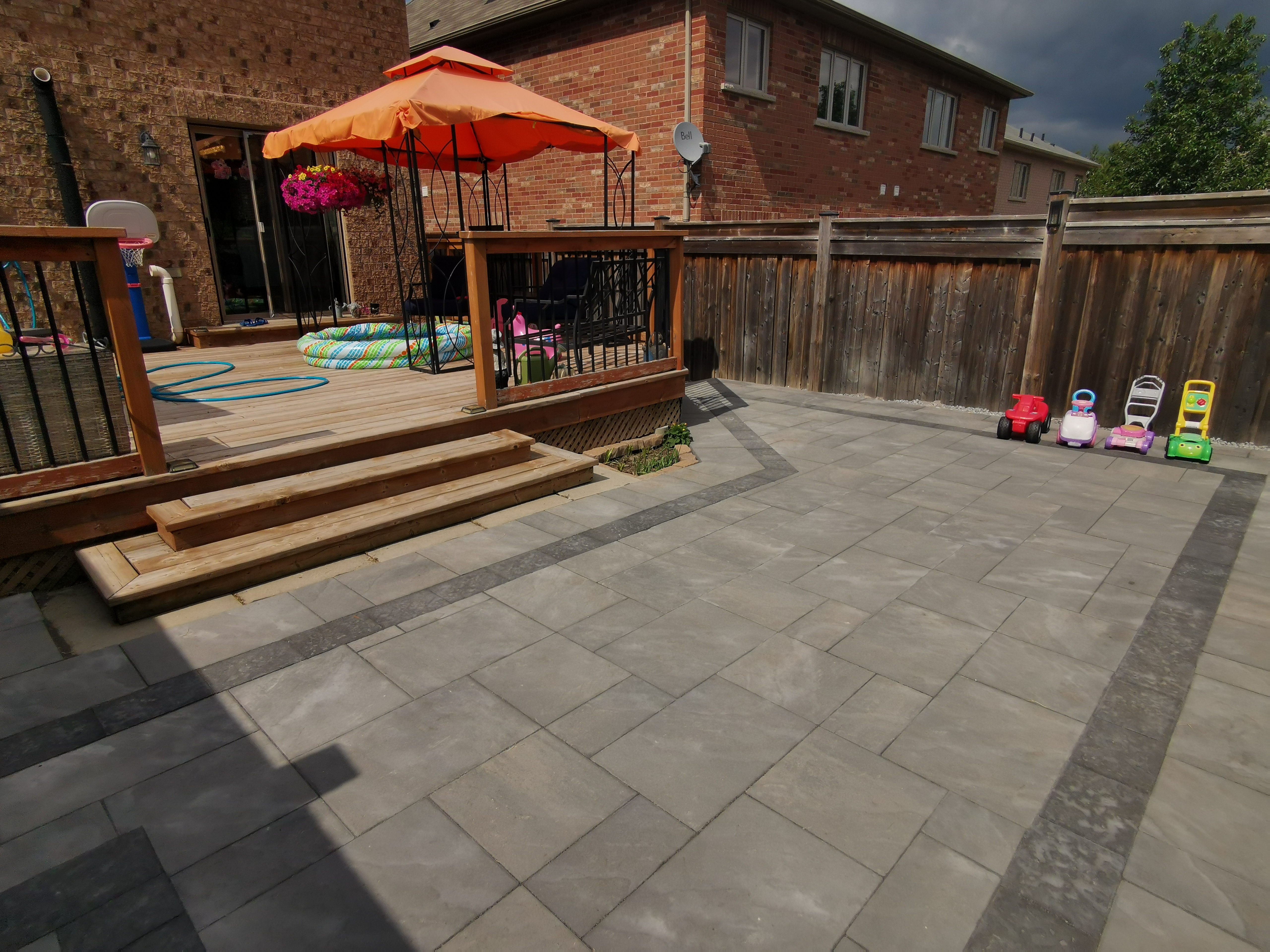 This Aurora backyard interlocking paver patio is designed to create a functional and inviting outdoor living space. Large-format concrete pavers are installed in a clean linear pattern with a contrasting border, enhancing visual definition and long-term durability. The patio seamlessly connects to a raised wooden deck with integrated steps, allowing smooth movement between indoor and outdoor areas. This professionally built interlock patio offers excellent drainage, stability, and low maintenance, making it ideal for family use and everyday enjoyment in residential backyards across Aurora.