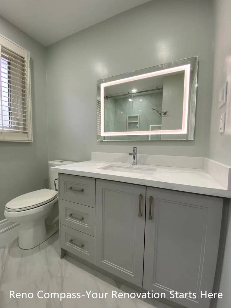 This Oakville interior renovation showcases a modern bathroom vanity designed for both functionality and clean aesthetics. The custom grey shaker-style vanity provides ample storage, paired with a quartz countertop for durability and easy maintenance. An LED backlit mirror enhances visibility while adding a contemporary touch to the space. Neutral wall tones and large-format floor tiles contribute to a bright, calm atmosphere, making the bathroom feel spacious and comfortable. This design emphasizes practical daily use while reflecting the quality craftsmanship and refined detailing delivered by a professional Toronto renovation company.