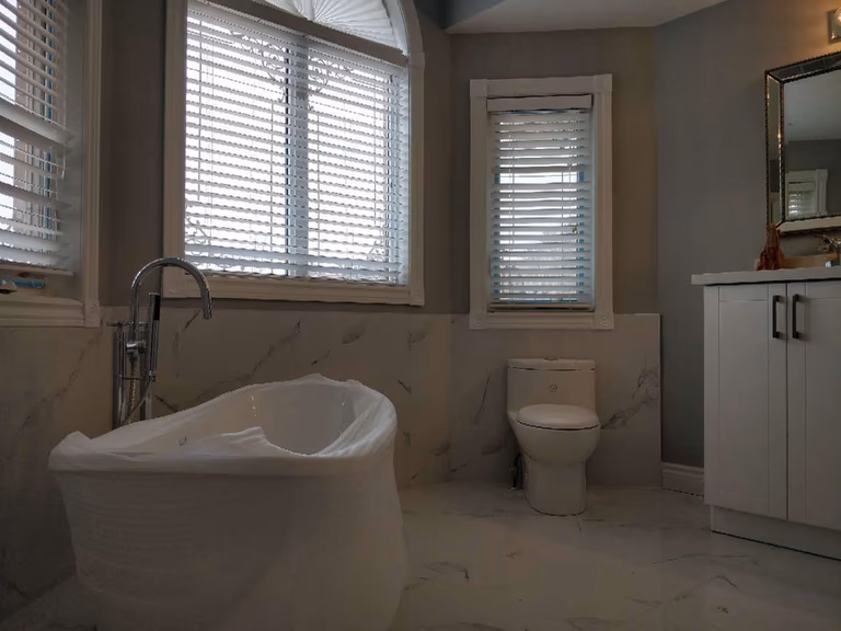 Luxury bathroom renovation in Ajax featuring a freestanding soaking bathtub, marble-style wall and floor tiles, modern white vanity, and large windows that bring natural light into the space. This elegant bathroom upgrade combines comfort and contemporary design, completed by a professional Toronto renovation company specializing in high-quality residential remodeling.