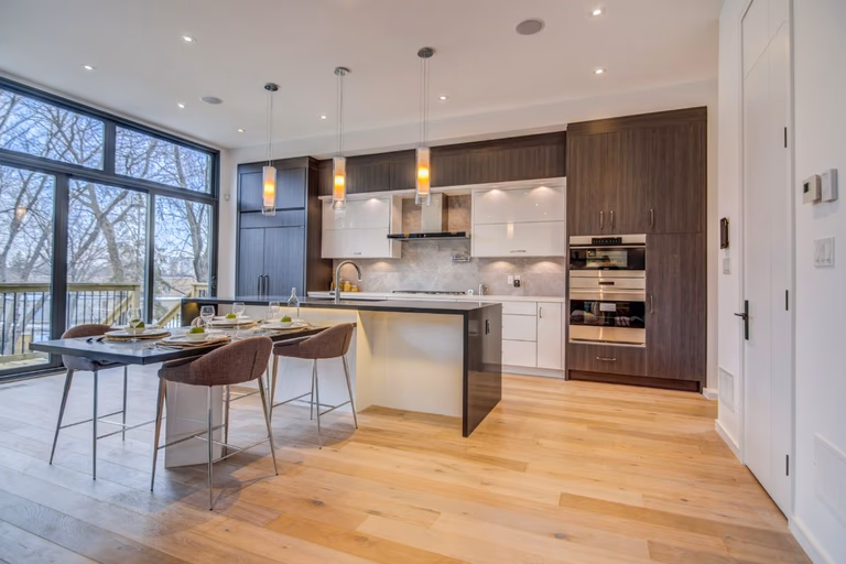 Contemporary Toronto kitchen renovation featuring custom two-tone cabinetry, large central island with seating, quartz countertops, and modern pendant lighting. Floor-to-ceiling windows bring in natural light, enhancing the bright open-concept layout. This modern kitchen was completed by a professional Toronto renovation company, combining functionality, storage, and elegant residential design for upscale living.