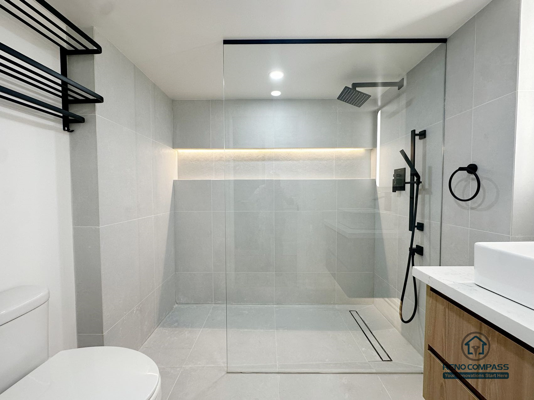 This bathroom renovation showcases a clean curbless shower layout with integrated wall niche lighting and a linear floor drain, creating a seamless and visually calm space. Large-format tiles, soft neutral tones, and matte black fixtures work together to achieve a balanced, modern look without visual clutter. The shower area is carefully planned to ensure proper drainage while maintaining a continuous floor surface, improving both daily comfort and long-term functionality.