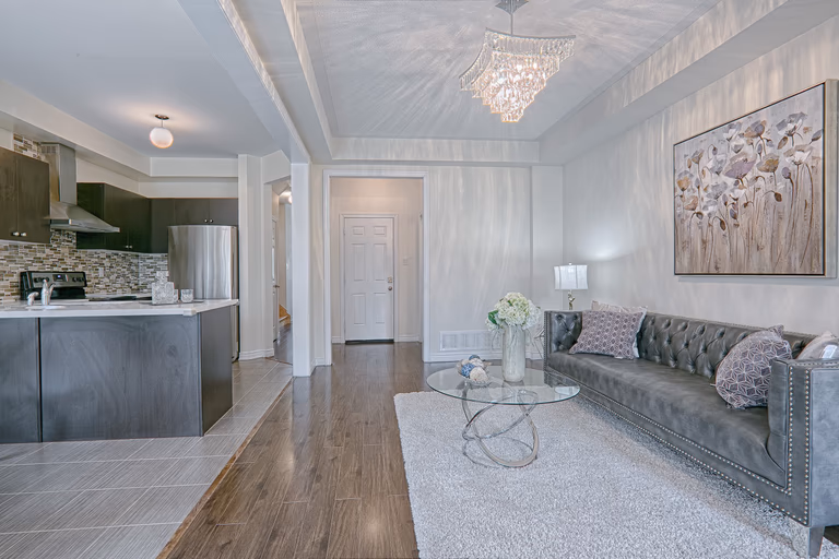 This contemporary chic interior features a refined open-concept living room with dark hardwood flooring, a textured area rug, and a statement crystal chandelier that casts elegant light reflections across the ceiling. The modern fireplace wall with mounted TV, large windows, and soft neutral wall tones create a bright yet cozy atmosphere. Styled with geometric side tables and luxury upholstery, this space reflects the craftsmanship and detail-oriented execution of a trusted Toronto renovation company specializing in modern interior transformations.