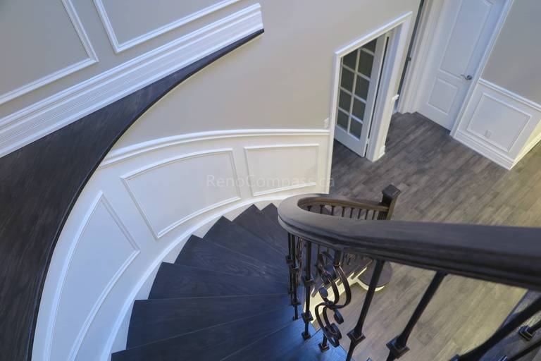 This Markham interior renovation features a stunning custom curved staircase with dark wood treads, white risers, and elegant wrought iron railings. Detailed wall panel molding and modern flooring enhance the sophisticated architectural design, creating a luxurious focal point within the home. Completed by Reno Compass, a professional renovation contractor specializing in custom staircases and high-end interior renovations across Markham and the Greater Toronto Area.