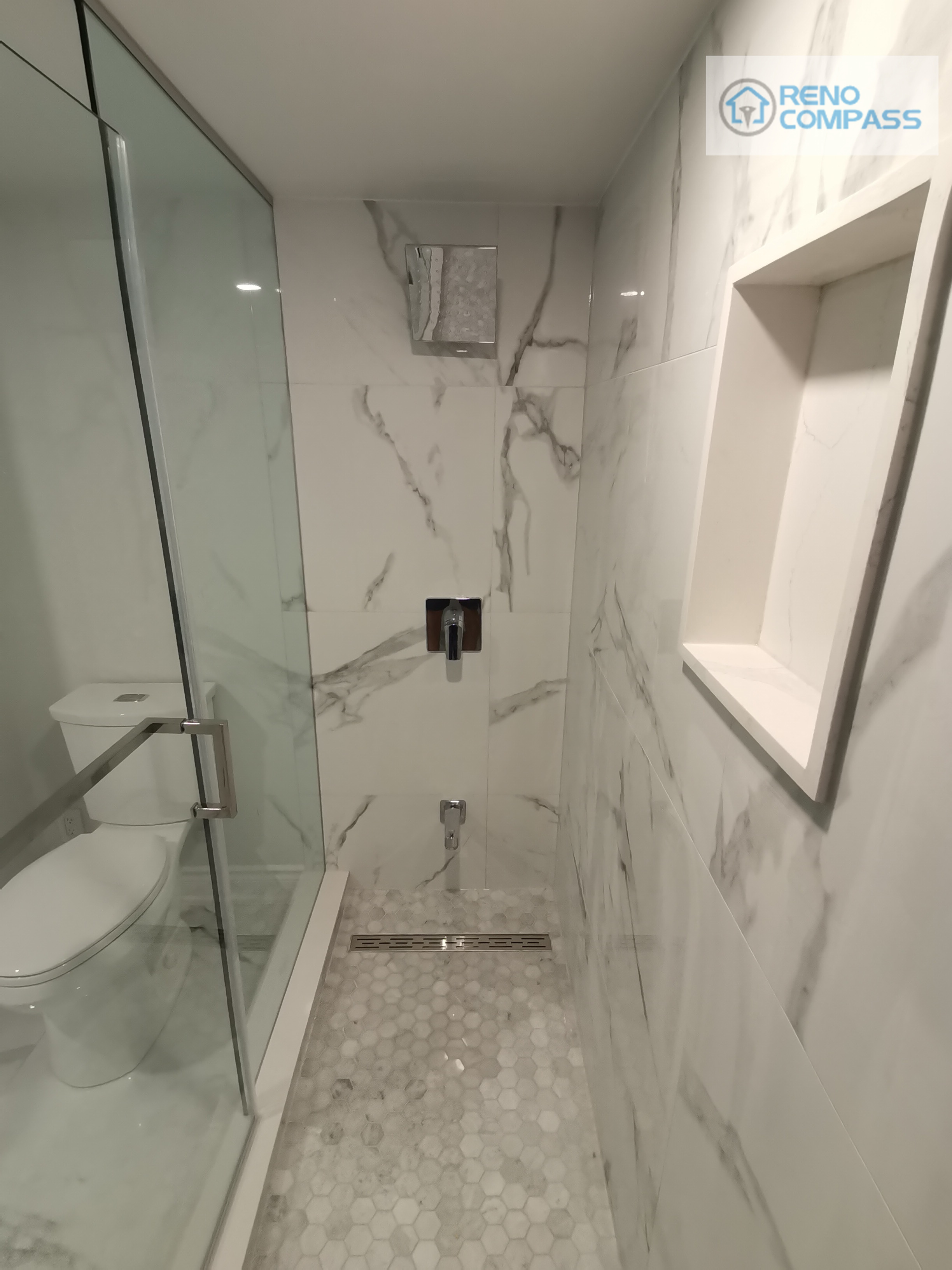 This walk-in shower from the Woodbridge basement renovation highlights a clean, modern layout with full-height marble-look wall tiles and a frameless glass enclosure. A built-in wall niche provides practical storage without interrupting the visual flow, while the mosaic shower floor improves slip resistance and drainage performance. Black shower fixtures add contrast and definition, creating a balanced and functional shower space suitable for everyday basement use or guest accommodation.