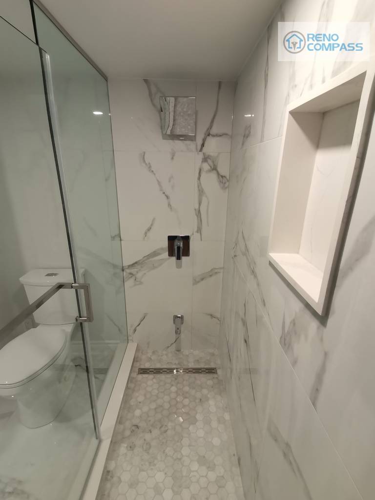 This walk-in shower from the Woodbridge basement renovation highlights a clean, modern layout with full-height marble-look wall tiles and a frameless glass enclosure. A built-in wall niche provides practical storage without interrupting the visual flow, while the mosaic shower floor improves slip resistance and drainage performance. Black shower fixtures add contrast and definition, creating a balanced and functional shower space suitable for everyday basement use or guest accommodation.