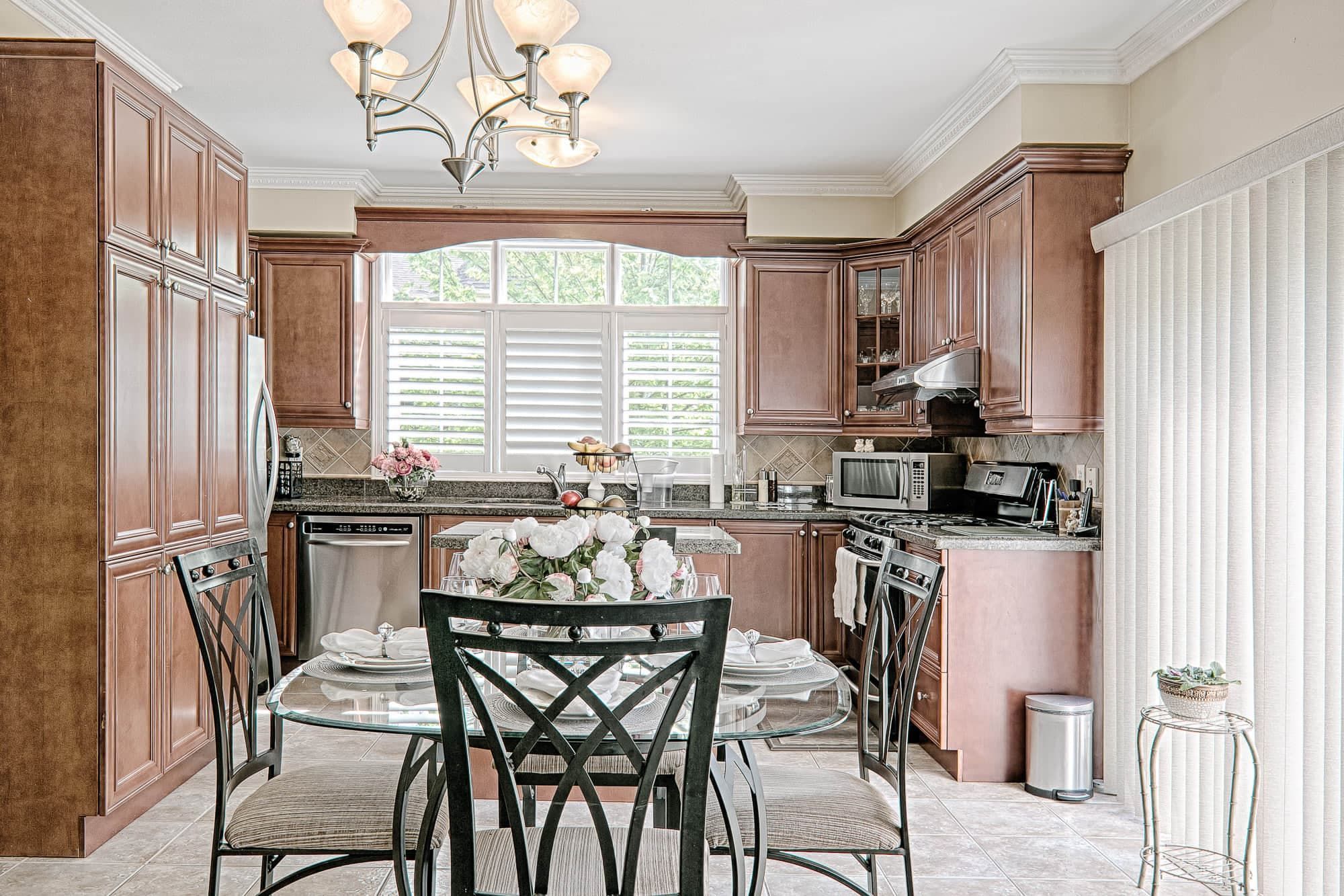 This bright traditional kitchen and dining area features rich wood cabinetry, granite countertops, and a cozy dining setup that creates a welcoming family environment. Large windows with plantation shutters allow natural light to illuminate the space, while the elegant chandelier and glass dining table enhance the overall warmth and sophistication. A beautiful example of functional kitchen renovation and classic home interior design inspiration for homeowners in Toronto and the GTA.