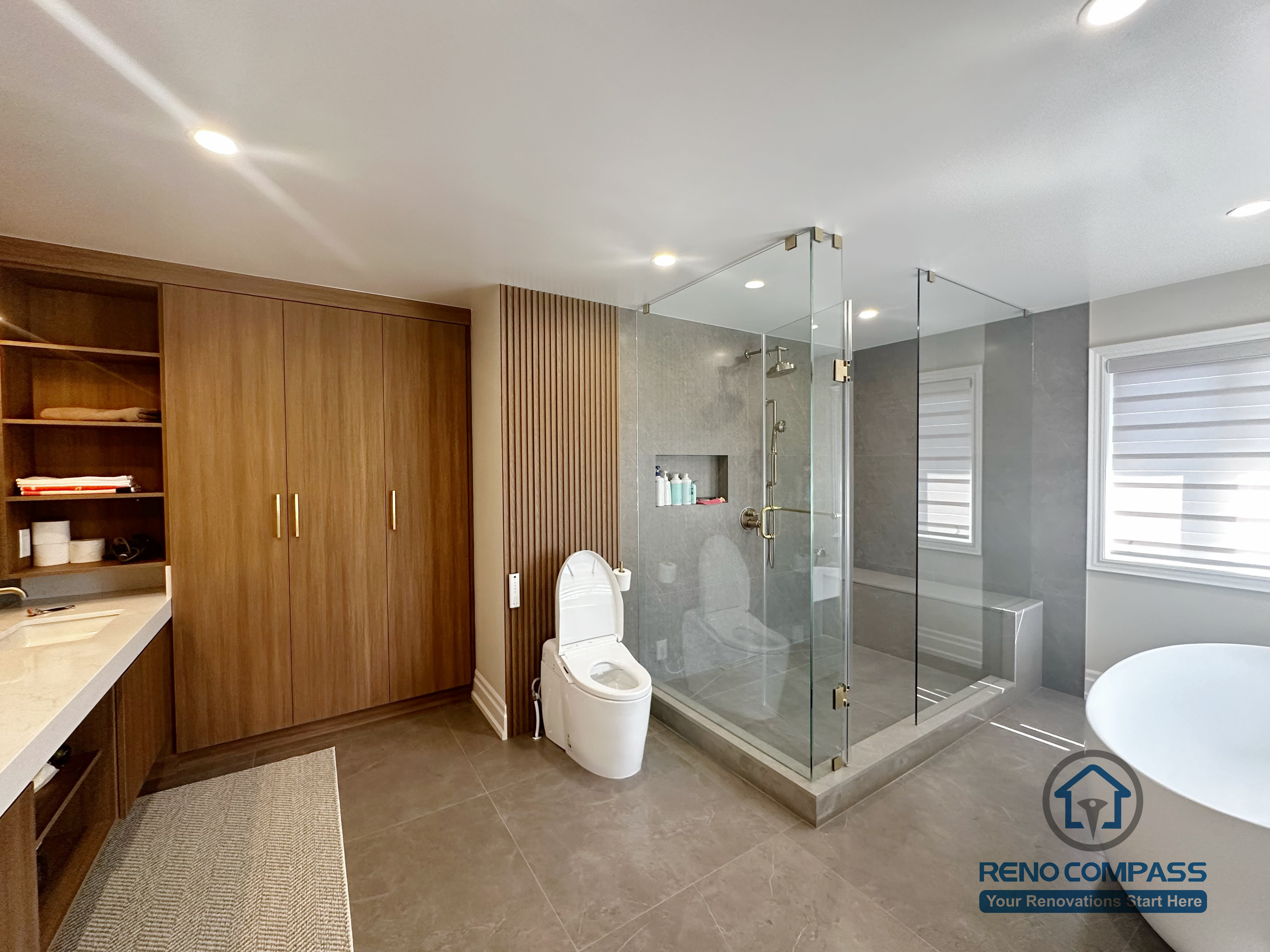 This image showcases a spacious primary bathroom renovation completed in North York, featuring a walk-in glass shower with built-in niche, a freestanding soaking tub, and custom wood storage cabinetry. Large-format floor tiles provide durability and visual continuity, while the frameless shower enclosure enhances openness and natural light. The layout balances functionality and comfort, creating a modern bathroom designed for everyday use and long-term living in North York homes.