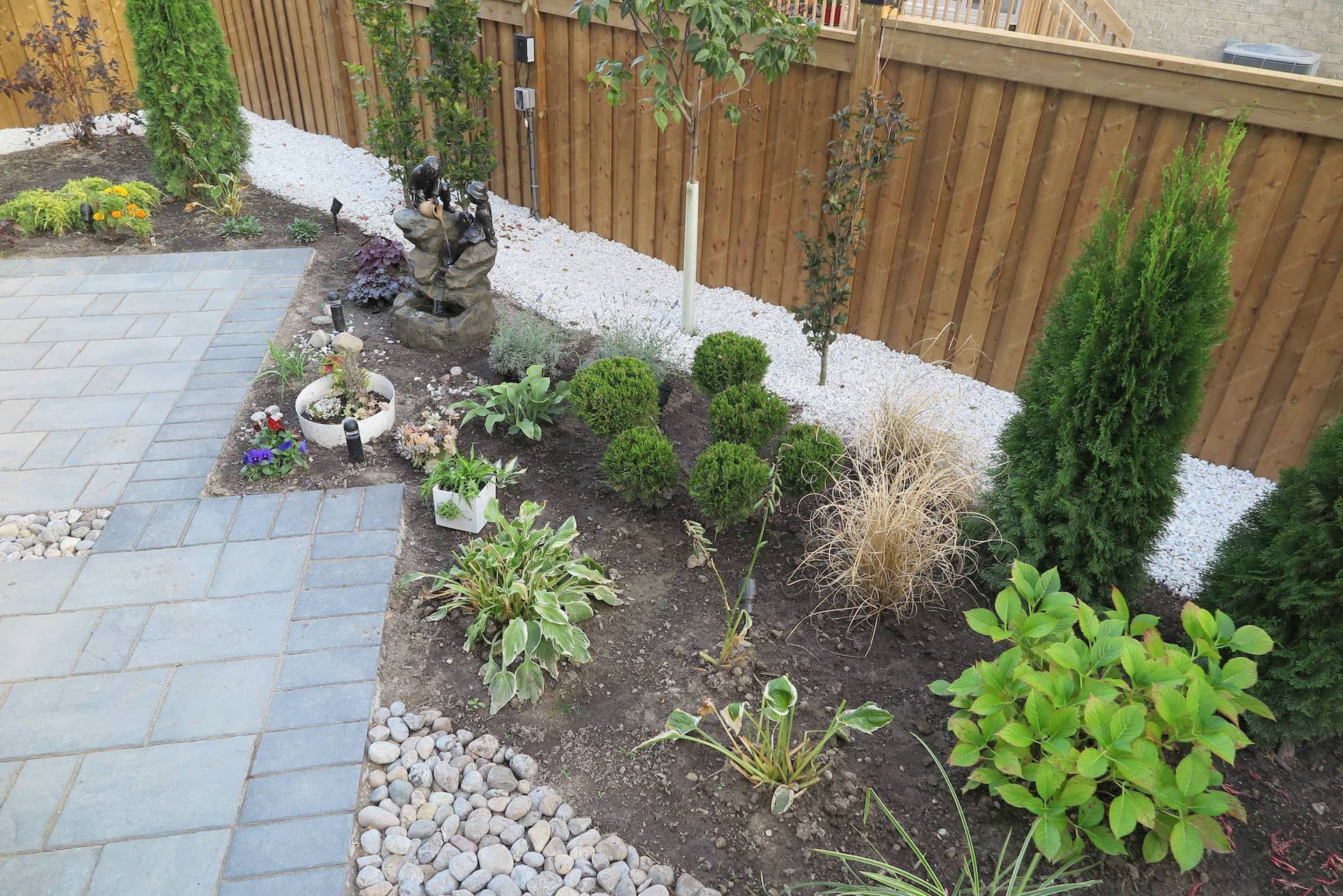 Backyard landscaping renovation in Vaughan featuring a modern interlocking patio, decorative river rock accents, layered garden beds, and a small ornamental water feature. This outdoor design blends hardscape and soft scape elements to create a relaxing backyard environment with strong visual balance and natural landscape texture.
