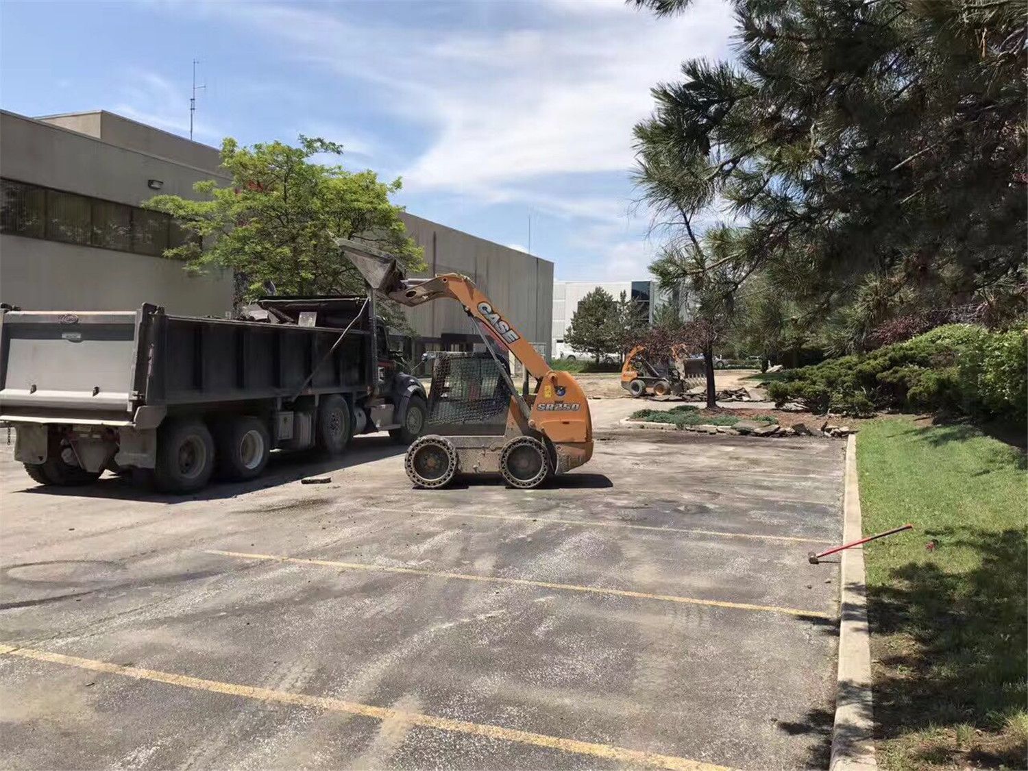 Toronto landscaping renovation project showing heavy equipment performing site preparation and excavation for a commercial parking area. The work includes asphalt removal, grading, and material hauling to prepare the site for new hardscape installation and improved landscape layout.