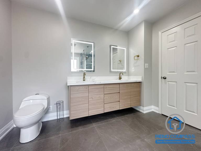 This Oakville bathroom renovation features a streamlined double vanity with integrated wood cabinetry, clean-lined countertops, and backlit mirrors for even task lighting. The design emphasizes practical storage, comfortable daily use, and visual balance, creating a calm and uncluttered bathroom environment. Completed by Reno Compass, this vanity layout reflects a thoughtful approach to modern Oakville bathroom design.