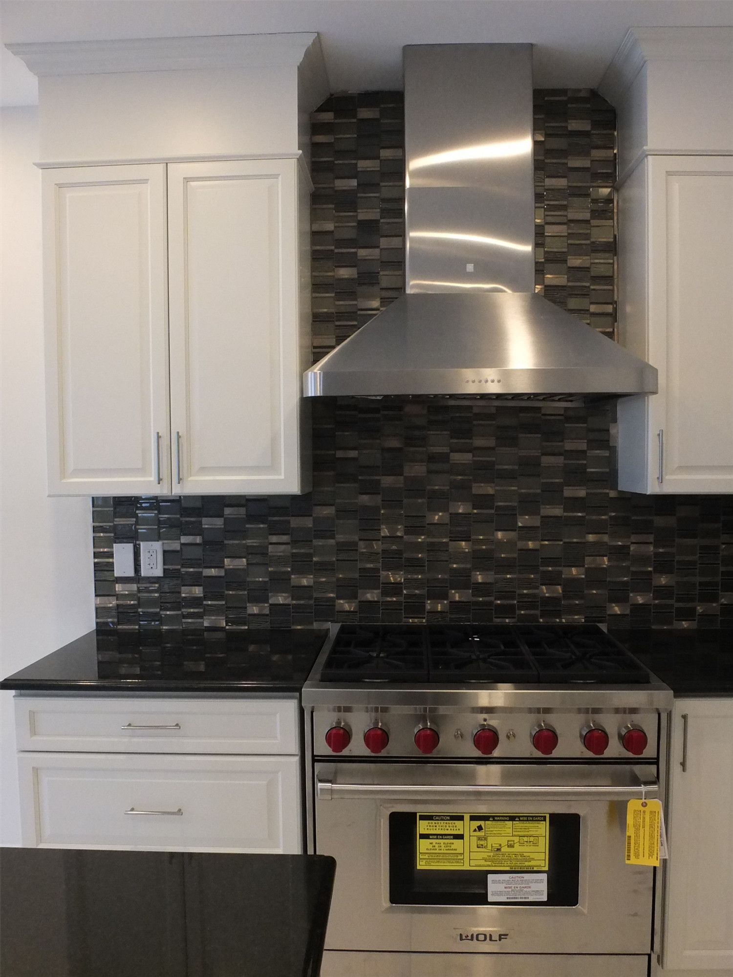 This Kleinburg interior renovation showcases a modern kitchen featuring custom white cabinetry, a spacious island with integrated sink, and sleek black countertops. The kitchen is highlighted by a professional stainless steel gas range and hood, complemented by a dark mosaic tile backsplash that adds depth and contrast. Elegant pendant lighting and large windows enhance the bright, open atmosphere, creating a refined and functional cooking space ideal for modern living.