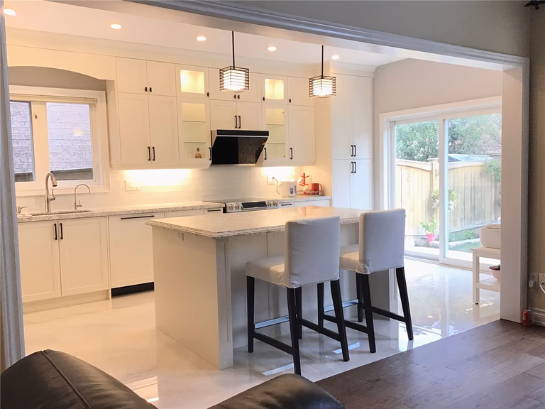 This Richmond Hill interior renovation features a bright open-concept kitchen centered around a large quartz island with seating. The space is designed with full-height custom cabinetry, clean white finishes, and modern pendant lighting that enhances both functionality and visual appeal. The symmetrical layout, under-cabinet lighting, and efficient workflow create a welcoming and practical environment for everyday living. Completed by a professional Toronto renovation company, this project reflects a refined and timeless kitchen design.