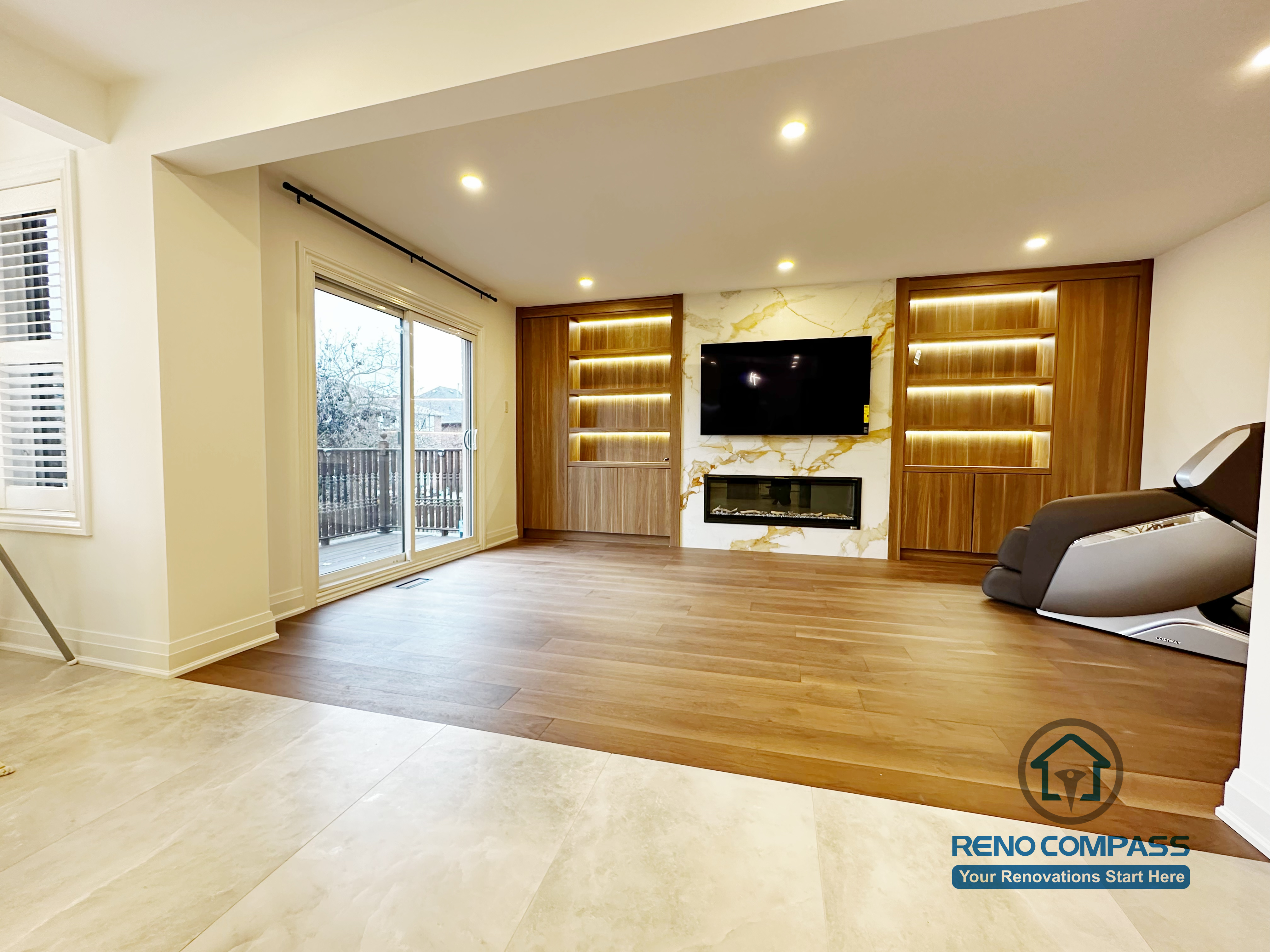 This image highlights a custom-built feature wall completed as part of a Markham interior renovation project. The design features warm wood paneling, integrated LED-lit display shelving, and a marble-look TV backdrop with a modern linear fireplace. Wide-plank hardwood flooring and recessed ceiling lights create a clean, open feel, while the built-in elements add both storage and visual focus. This space was designed to seamlessly connect with the adjacent kitchen and dining areas, reflecting the cohesive craftsmanship of a professional Toronto renovation company.