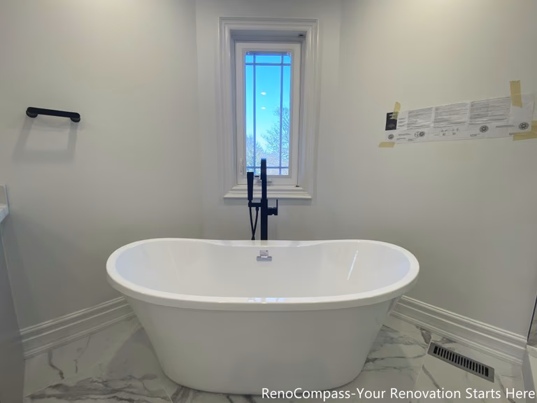 This Thornhill bathroom renovation features a freestanding bathtub positioned beneath a window, creating a calm and balanced focal point within the space. The tub is paired with a matte black floor-mounted faucet, adding a modern contrast to the light marble-look floor tiles. Natural daylight enhances the clean, minimal design, while the open layout promotes comfort, relaxation, and long-term functionality for everyday use.