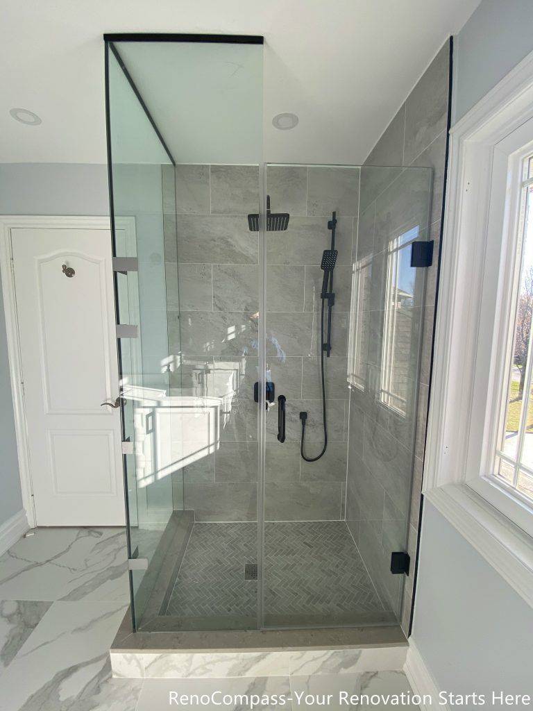 This Thornhill bathroom renovation features a modern walk-in shower enclosed with frameless glass panels, creating a bright and visually open space. The shower is finished with large-format gray wall tiles, a patterned shower base, and matte black fixtures for a clean, contemporary contrast. Natural light from the window enhances the overall brightness, while the glass enclosure replaces traditional drywall to significantly improve spatial flow, functionality, and long-term usability.