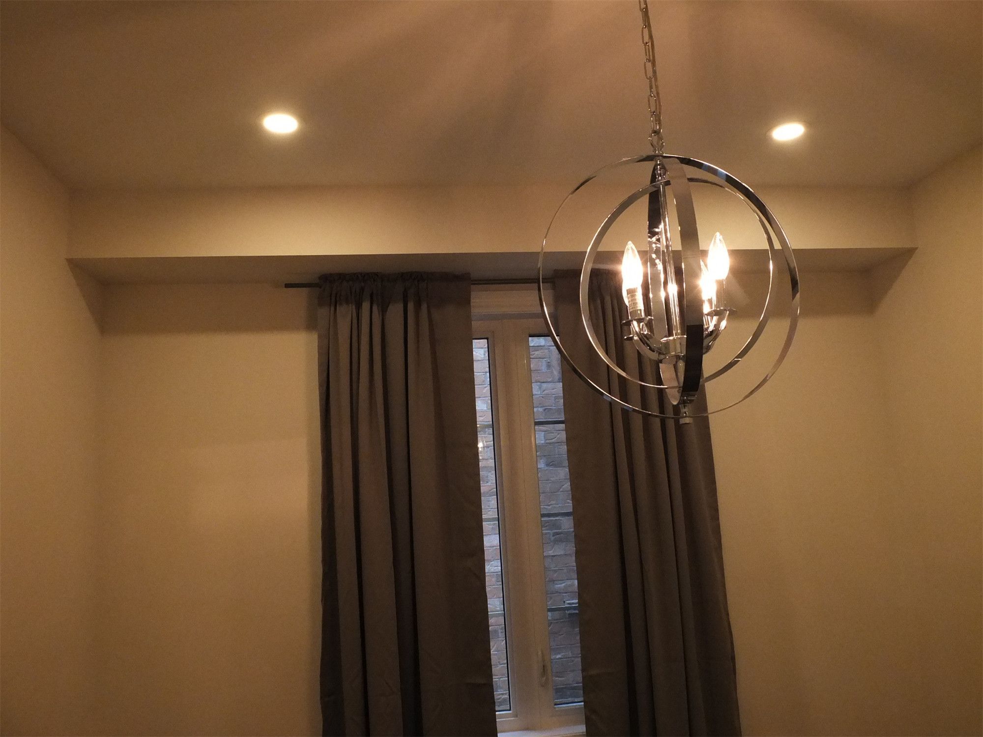 This Kleinburg interior renovation highlights a modern chandelier suspended above the living space, complemented by recessed pot lights and natural light from the window. The contemporary lighting fixture adds a decorative focal point while providing warm ambient illumination. Combined with soft wall finishes and custom window treatments, the design enhances the comfort and elegance of the renovated interior.