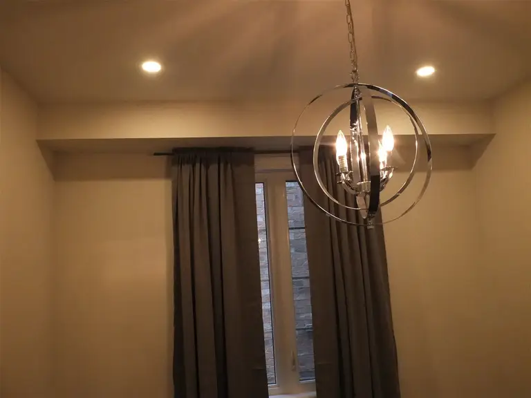 This Kleinburg interior renovation highlights a modern chandelier suspended above the living space, complemented by recessed pot lights and natural light from the window. The contemporary lighting fixture adds a decorative focal point while providing warm ambient illumination. Combined with soft wall finishes and custom window treatments, the design enhances the comfort and elegance of the renovated interior.