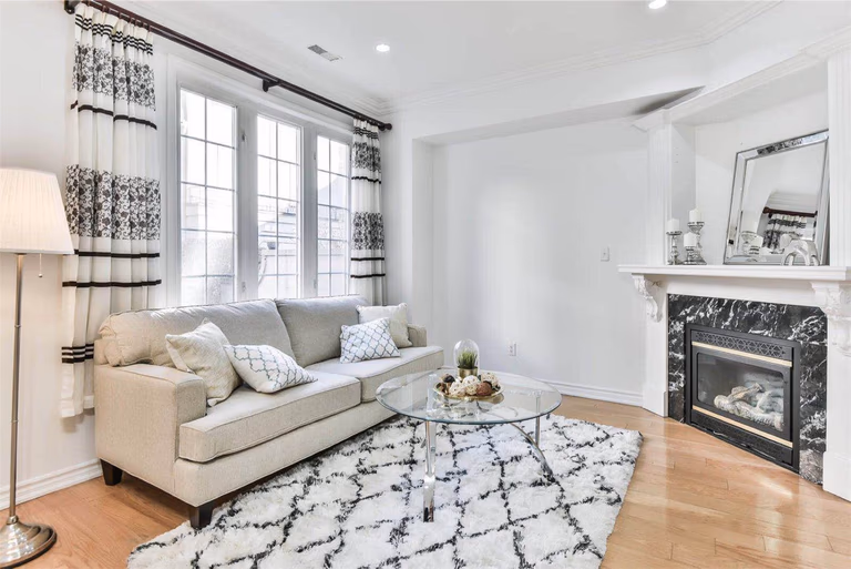 This project features a professionally staged living room in a Toronto townhouse prepared for sale. The space highlights a bright and welcoming atmosphere with a light-colored sofa, patterned curtains, soft textured rug, and a modern glass coffee table. Natural light from large windows enhances the clean white interior, while the fireplace adds warmth and elegance to the room. Professional home staging helps present the property at its best, allowing potential buyers to better visualize the space and increasing overall market appeal.