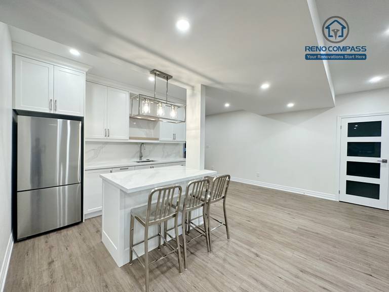 This Bradford basement renovation transforms the lower level into a bright, open-concept living and entertainment space. The layout integrates a custom wet bar with full-height cabinetry, quartz countertop, and built-in sink, creating a functional transition between the entertainment zone and seating area. Recessed LED lighting throughout the ceiling enhances brightness and eliminates the typical basement dark feel, while durable wood-look flooring provides warmth and continuity across the space. Structural columns were carefully incorporated into the design to maintain openness without compromising support. This basement renovation reflects Reno Compass’ approach to practical space planning, clean finishes, and long-term usability for modern family living.