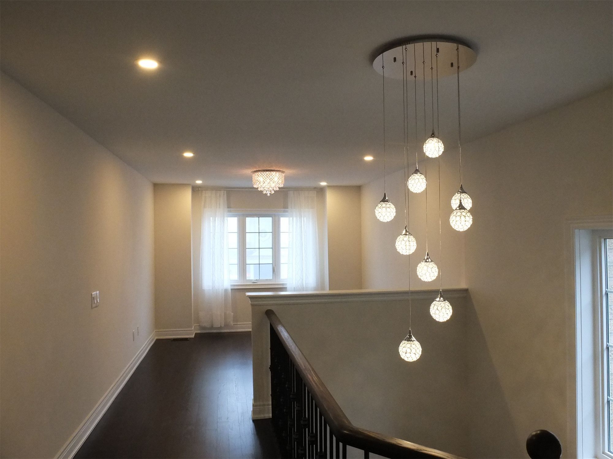 This Kleinburg interior renovation features a dramatic multi-pendant chandelier installed above the staircase, creating a striking focal point for the upper hallway. The cascading glass lights add elegance and depth to the open stairwell, while recessed pot lights provide balanced ambient lighting. Paired with dark hardwood flooring, neutral walls, and natural daylight from nearby windows, the space feels bright, modern, and architecturally refined.