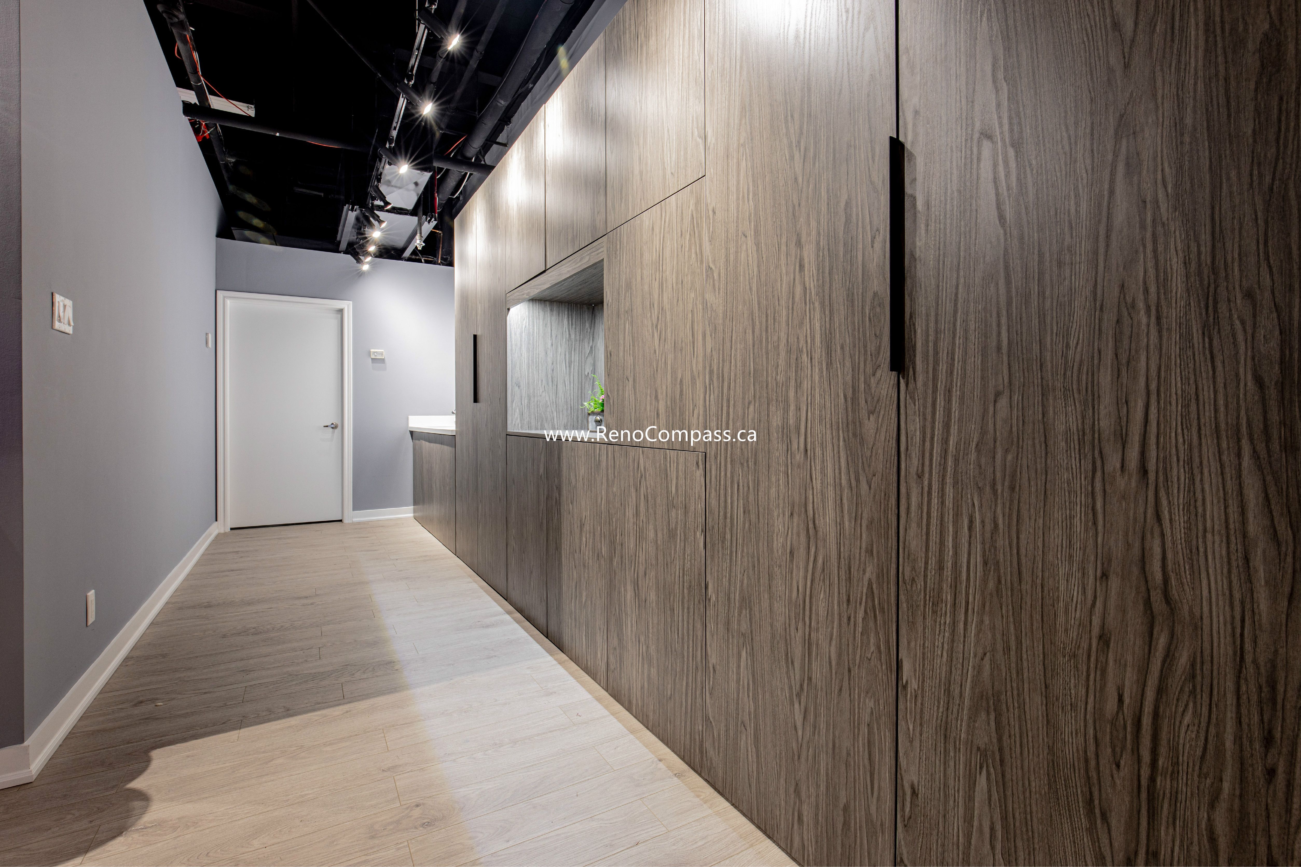 High-end custom wood cabinetry and integrated millwork for a commercial hallway. Features modern track lighting, exposed black ceilings, and luxury vinyl flooring. A professional interior renovation project by RenoCompass in Richmond Hill.