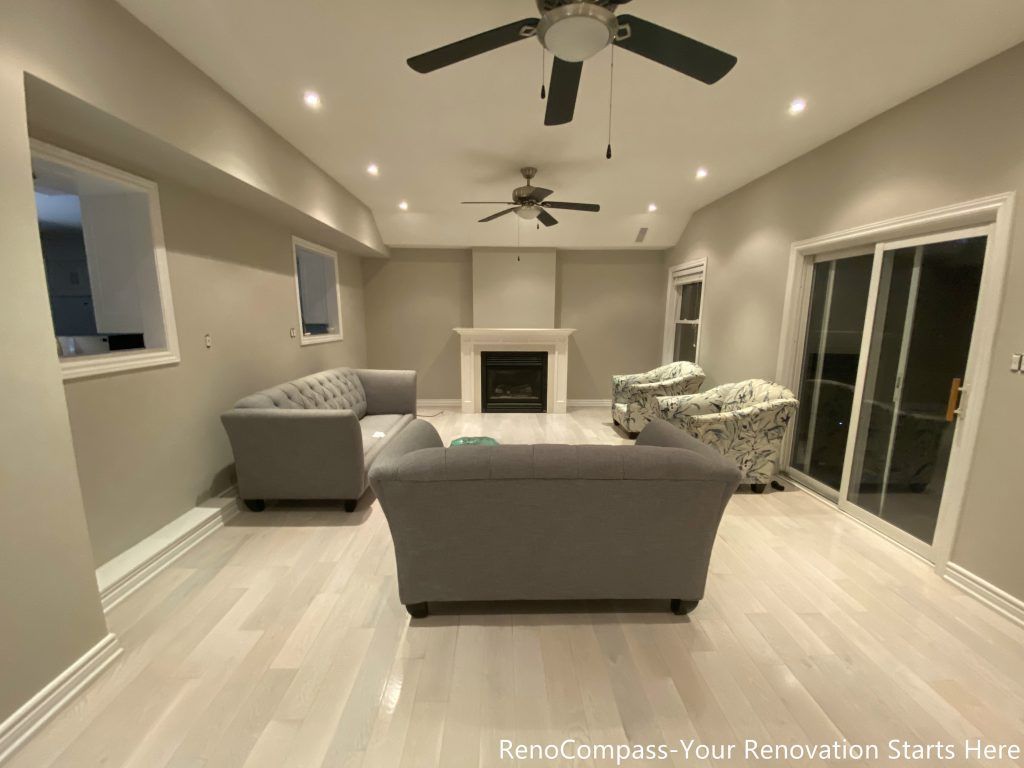 This completed living room renovation features light wood flooring, a central fireplace, and a well-balanced recessed lighting layout that enhances both comfort and spatial depth. Ceiling fans and large windows improve airflow and natural light, creating a relaxed and functional environment for everyday living and family gathering. Designed and completed by Reno Compass, this space reflects a clean, timeless interior foundation built for long-term use.