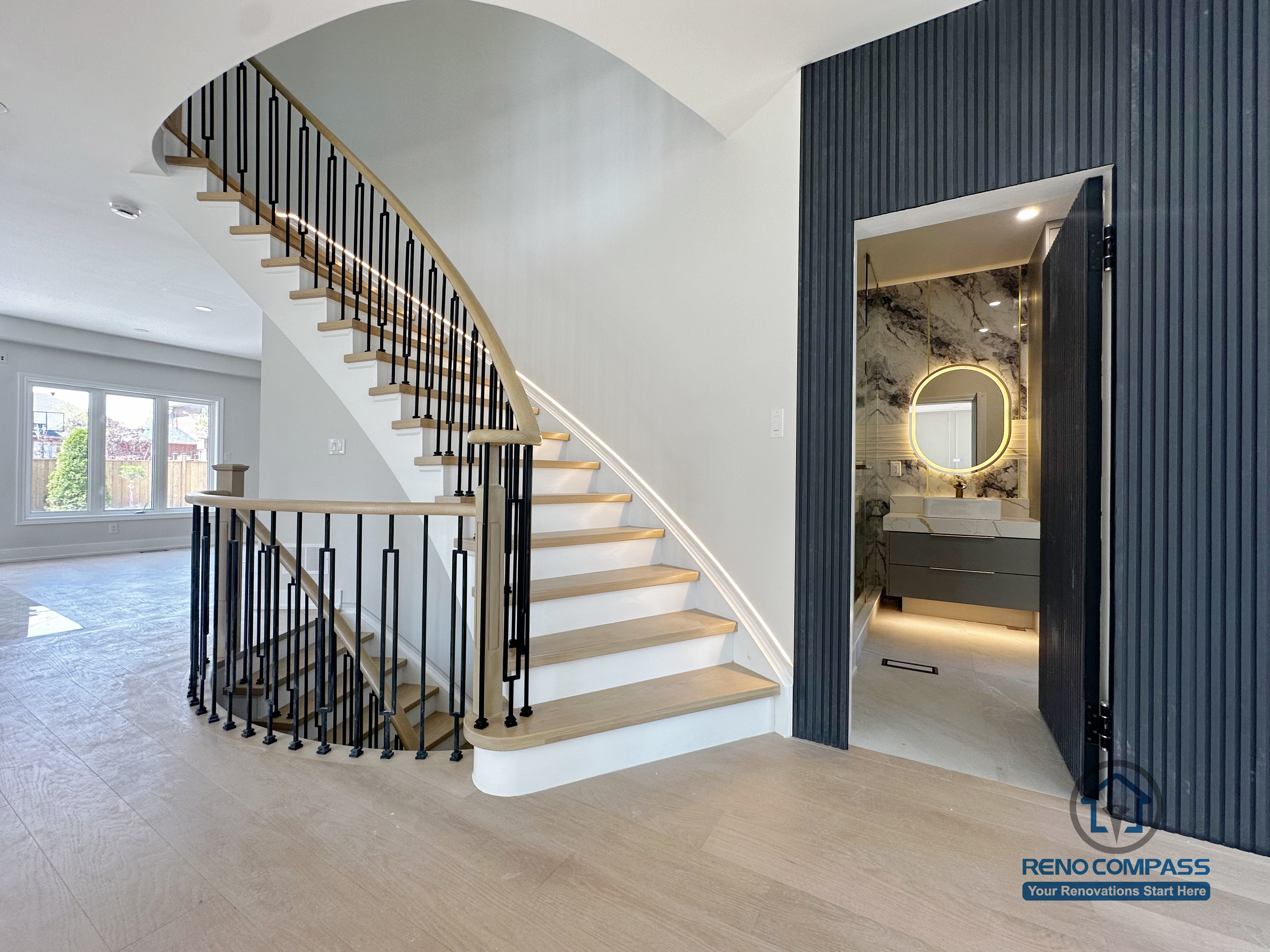 This image captures a refined transition space from a Markham interior renovation, featuring a curved staircase, architectural feature wall, and a discreet powder room entrance. The staircase design combines light wood treads with black metal railings, creating a clean contrast while maintaining visual openness throughout the main floor.

The curved stair profile softens the overall geometry of the space and enhances circulation flow between levels. Adjacent to the staircase, a vertical slat feature wall in a dark finish adds depth and definition, subtly framing the entry to the powder room.

Inside the powder room, layered lighting and a round illuminated mirror introduce a sense of intimacy and refinement, complementing the modern material palette used throughout the home. This design approach reflects Reno Compass’s focus on spatial continuity—where staircases, entry zones, and functional rooms are seamlessly integrated into a cohesive interior renovation in