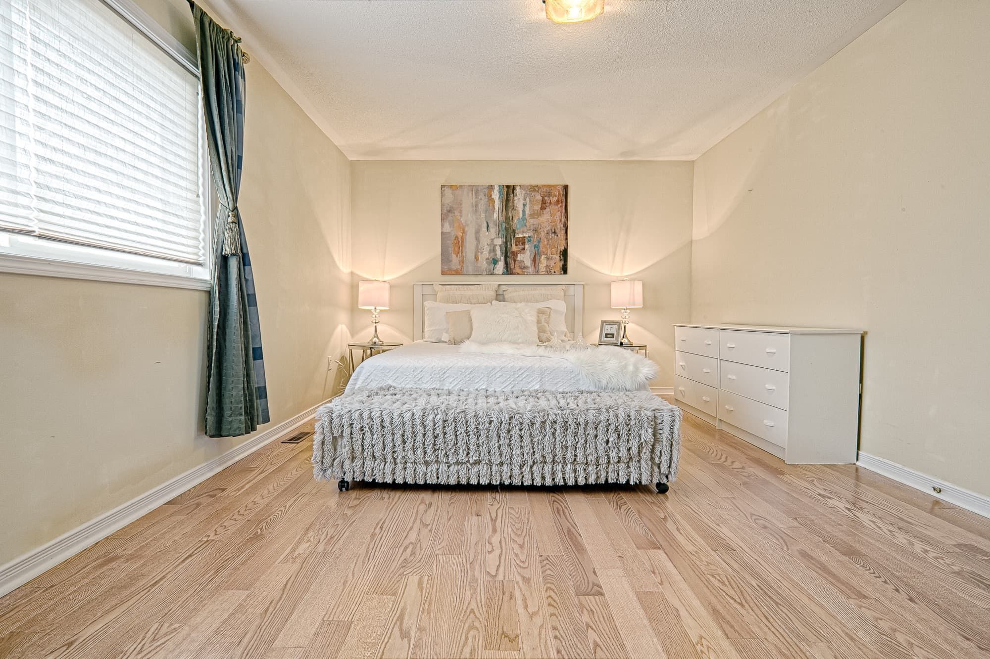 This spacious master bedroom features a simple and lovely interior design with natural hardwood flooring, soft neutral wall tones, and a well-balanced furniture layout. Symmetrical bedside lighting and minimal decor create a clean, relaxing atmosphere, while large windows provide ample natural light. The design focuses on comfort, openness, and timeless style, making it suitable for family homes and long-term residential living.