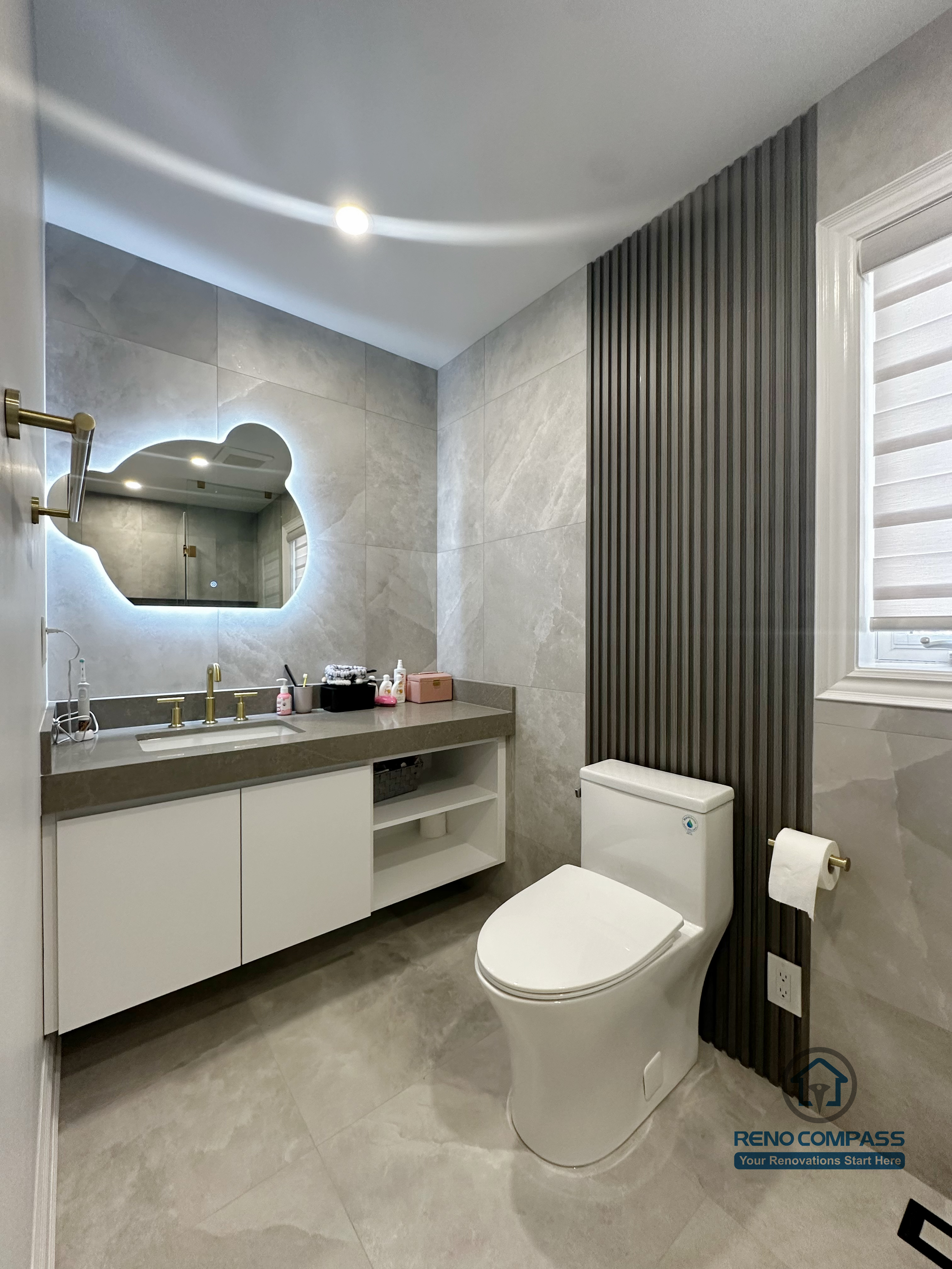 This image showcases the vanity and toilet area of a North York bathroom renovation, featuring a floating white vanity paired with a quartz countertop for a clean, modern look. The sculptural backlit mirror adds soft ambient lighting while enhancing depth and visual interest. Large-format grey tiles create a seamless backdrop, complemented by a vertical slat accent wall that introduces texture and contrast. The layout prioritizes functionality, storage efficiency, and a refined contemporary aesthetic suitable for everyday use.