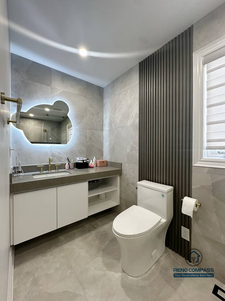 This image showcases the vanity and toilet area of a North York bathroom renovation, featuring a floating white vanity paired with a quartz countertop for a clean, modern look. The sculptural backlit mirror adds soft ambient lighting while enhancing depth and visual interest. Large-format grey tiles create a seamless backdrop, complemented by a vertical slat accent wall that introduces texture and contrast. The layout prioritizes functionality, storage efficiency, and a refined contemporary aesthetic suitable for everyday use.