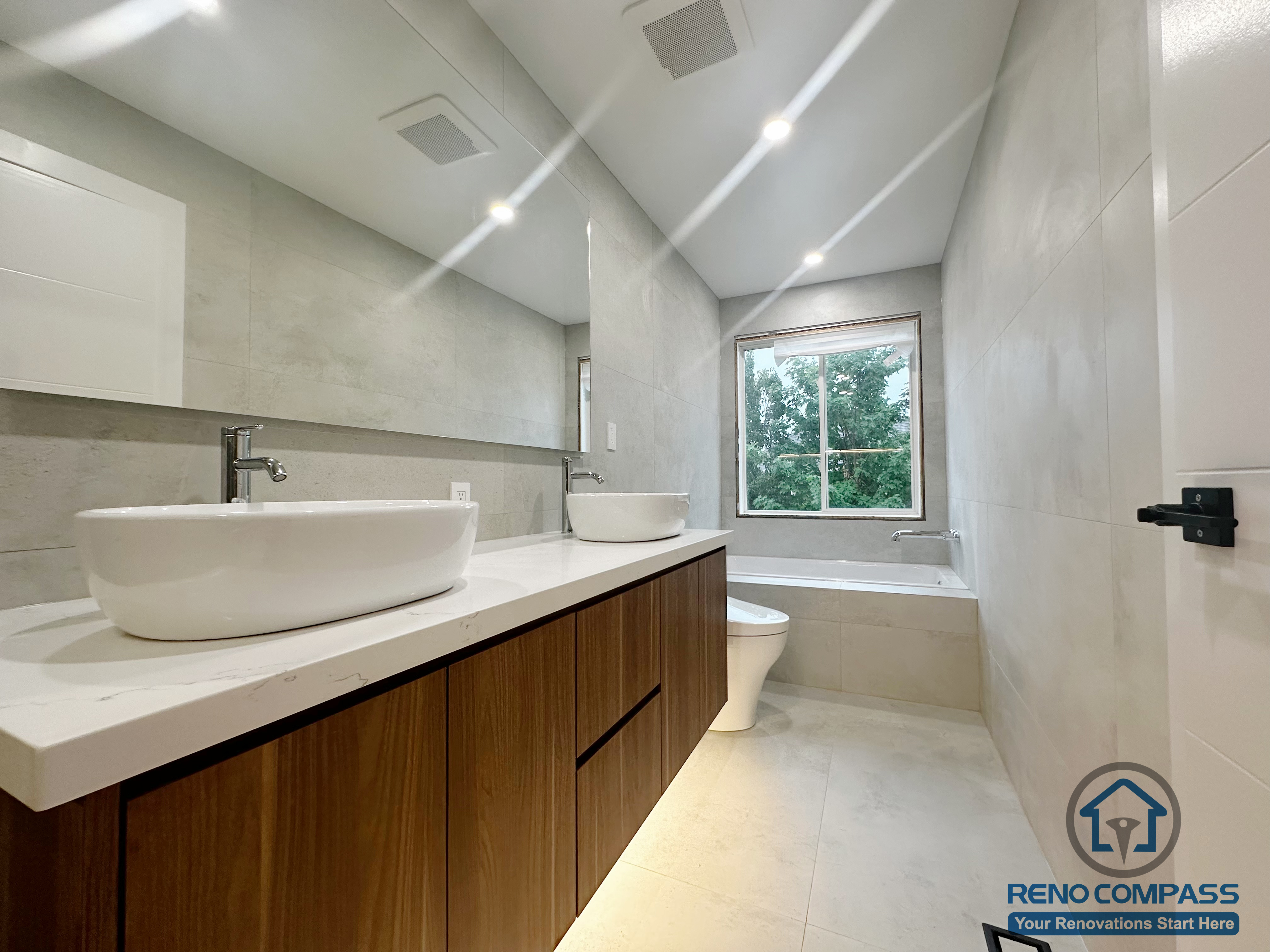 This Oakville bathroom renovation features a custom double vessel vanity with warm wood cabinetry, a quartz countertop, and integrated under-cabinet lighting. The space is finished with large-format wall tiles, a full-height mirror, and a clean, minimalist layout that maximizes natural light and everyday functionality. Designed and completed by Reno Compass, this bathroom balances modern aesthetics with long-term durability for residential use.