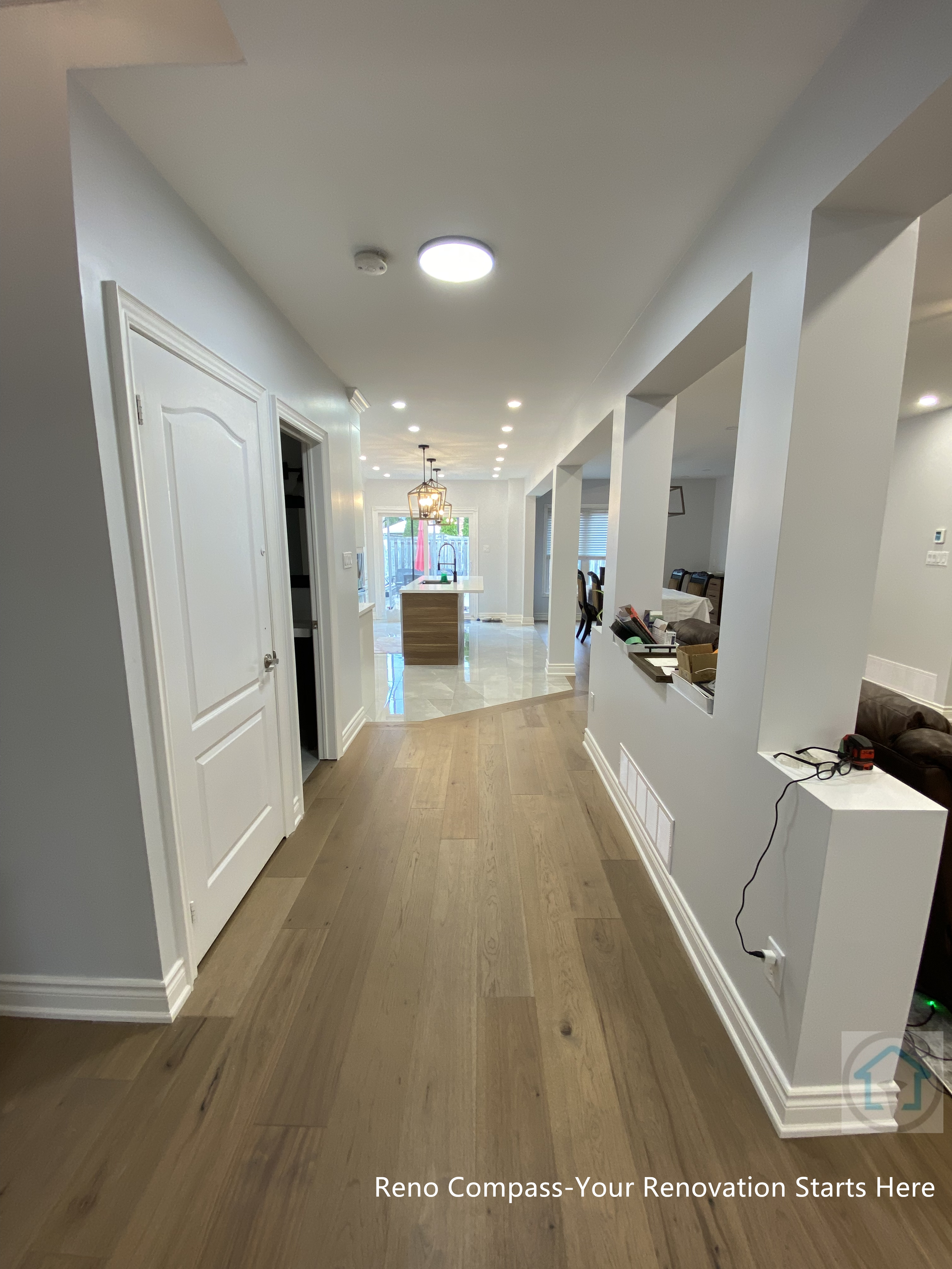 This image showcases a Newmarket interior renovation featuring a long, clean-lined hallway that connects the main living areas with the kitchen and dining space. The continuous hardwood flooring enhances visual flow and guides movement through the home, while recessed ceiling lights provide even, glare-free illumination. Open wall cut-outs along the right side improve sightlines and allow natural light to travel deeper into the interior, reinforcing the open-concept design. Neutral wall tones and simple trim details keep the space bright, modern, and timeless. Completed by a Toronto renovation company, this hallway renovation demonstrates thoughtful spatial planning, improved circulation, and a seamless transition between functional zones within the home.
