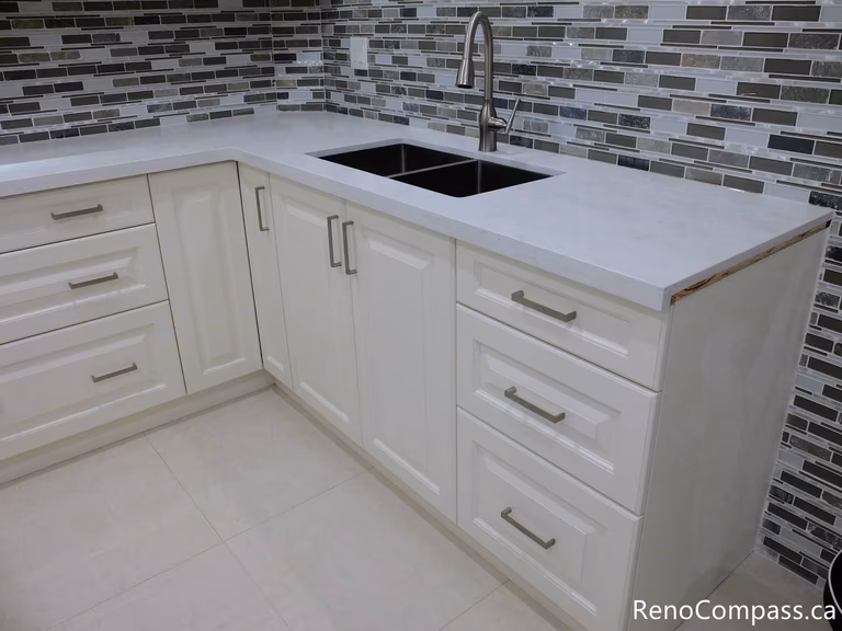 This Markham basement kitchen detail highlights a clean quartz countertop paired with an undermount sink and modern faucet. The white shaker cabinets and mosaic tile backsplash create a cohesive and contemporary look, while the practical layout ensures durability and ease of maintenance for everyday use. Completed by a trusted Toronto Renovation Company, Reno Compass Inc.