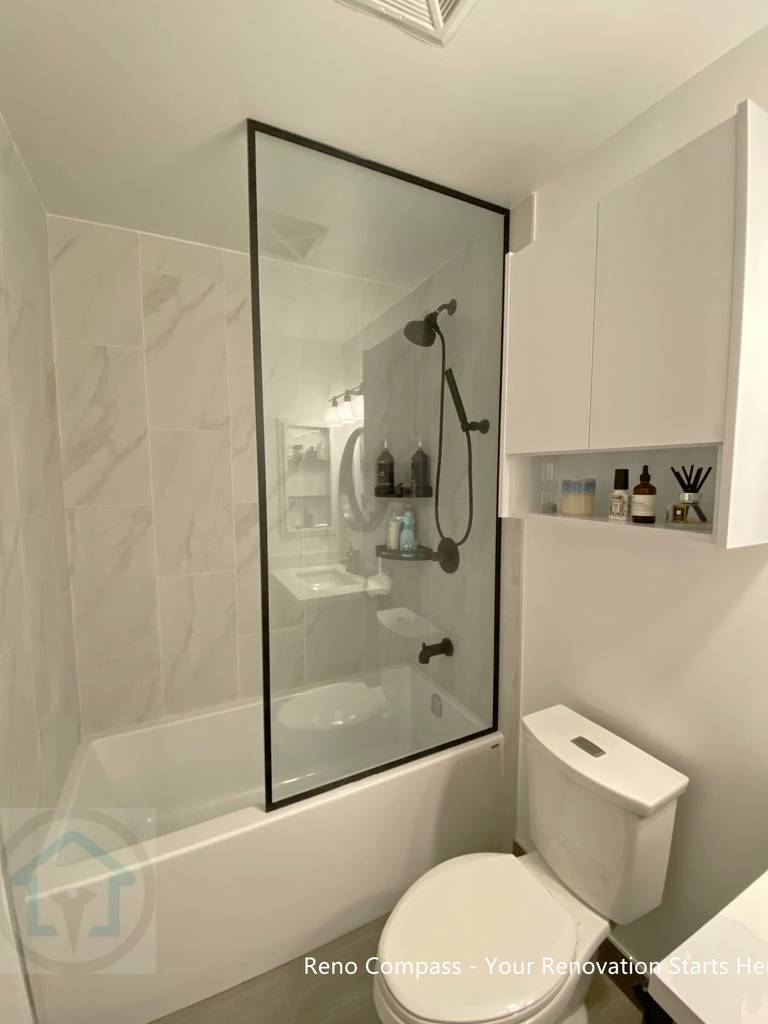 This North York condo bathroom renovation features a clean tub and shower combination with a custom black-framed glass panel, marble-look wall tiles, and matte black fixtures. Built-in wall storage improves daily usability while maintaining a streamlined, modern aesthetic ideal for compact condo bathrooms.