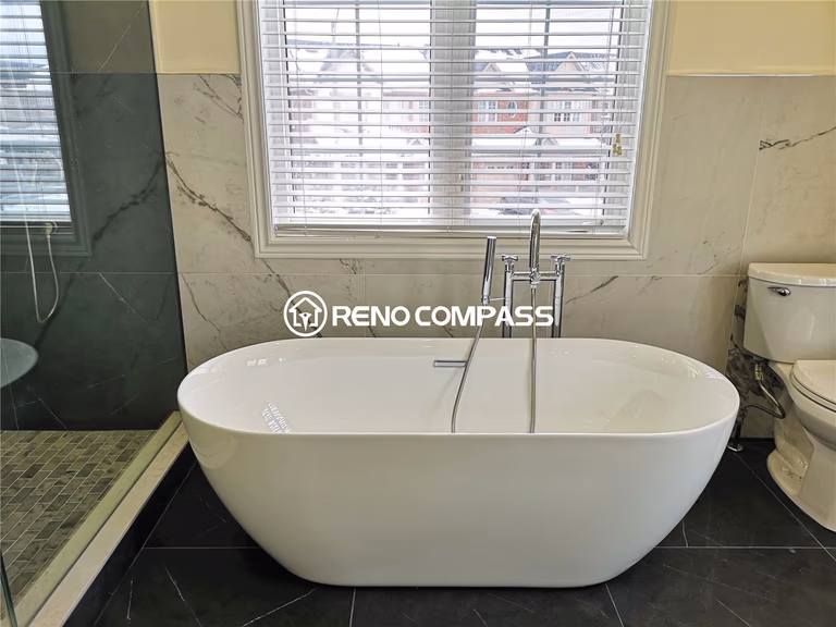 This luxurious Toronto bathroom renovation highlights a contemporary freestanding bathtub placed beside a large window, offering both comfort and elegance. The sleek marble wall, dark tile flooring, and chrome fixtures create a spa-inspired atmosphere. Designed by Reno Compass, this space captures modern sophistication and timeless design.