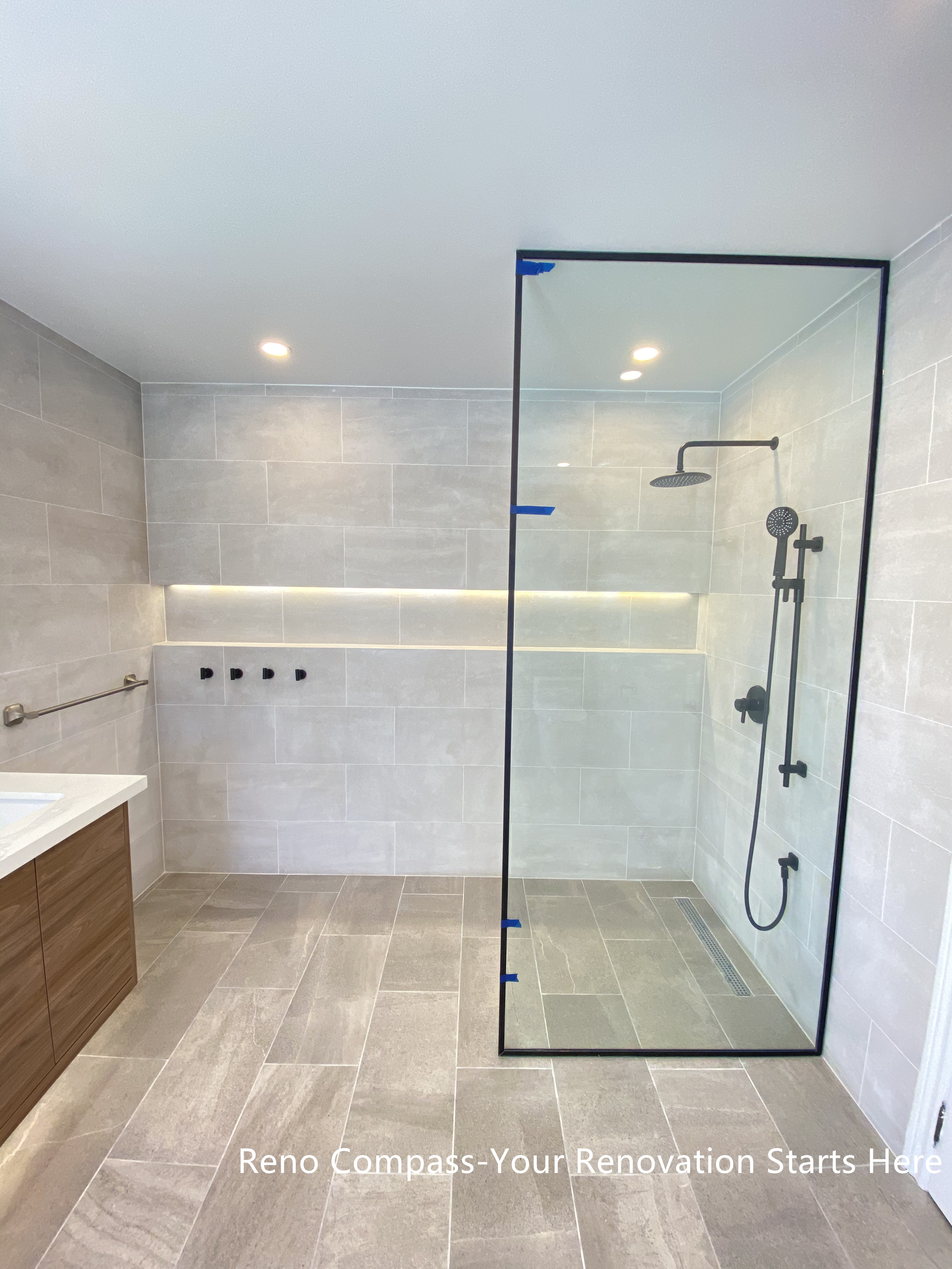 This Aurora bathroom renovation features a modern walk-in shower with a frameless glass enclosure, large-format wall tiles, and integrated niche lighting for both function and visual clarity. Matte black shower fixtures create a subtle contrast against the light tile palette, while the clean glass design maintains openness and easy circulation within the space. Recessed ceiling lighting and carefully aligned finishes contribute to a bright, refined shower environment. Designed and completed by Reno Compass, this bathroom renovation emphasizes practicality, clean detailing, and long-term usability.