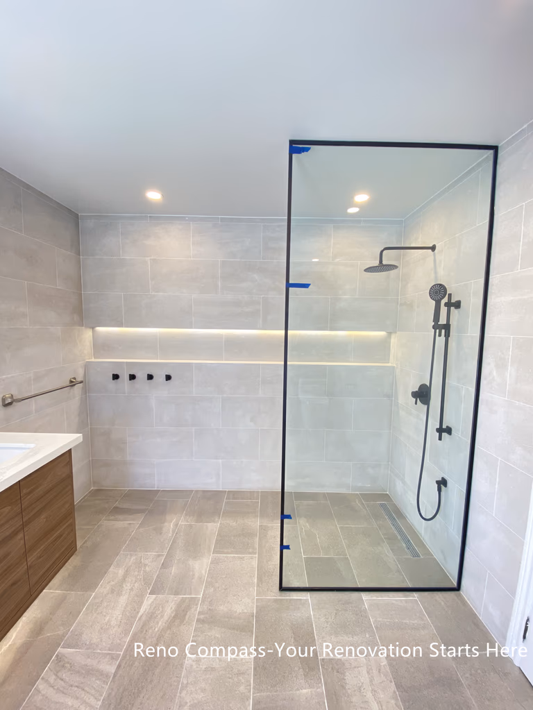 This Aurora bathroom renovation features a modern walk-in shower with a frameless glass enclosure, large-format wall tiles, and integrated niche lighting for both function and visual clarity. Matte black shower fixtures create a subtle contrast against the light tile palette, while the clean glass design maintains openness and easy circulation within the space. Recessed ceiling lighting and carefully aligned finishes contribute to a bright, refined shower environment. Designed and completed by Reno Compass, this bathroom renovation emphasizes practicality, clean detailing, and long-term usability.