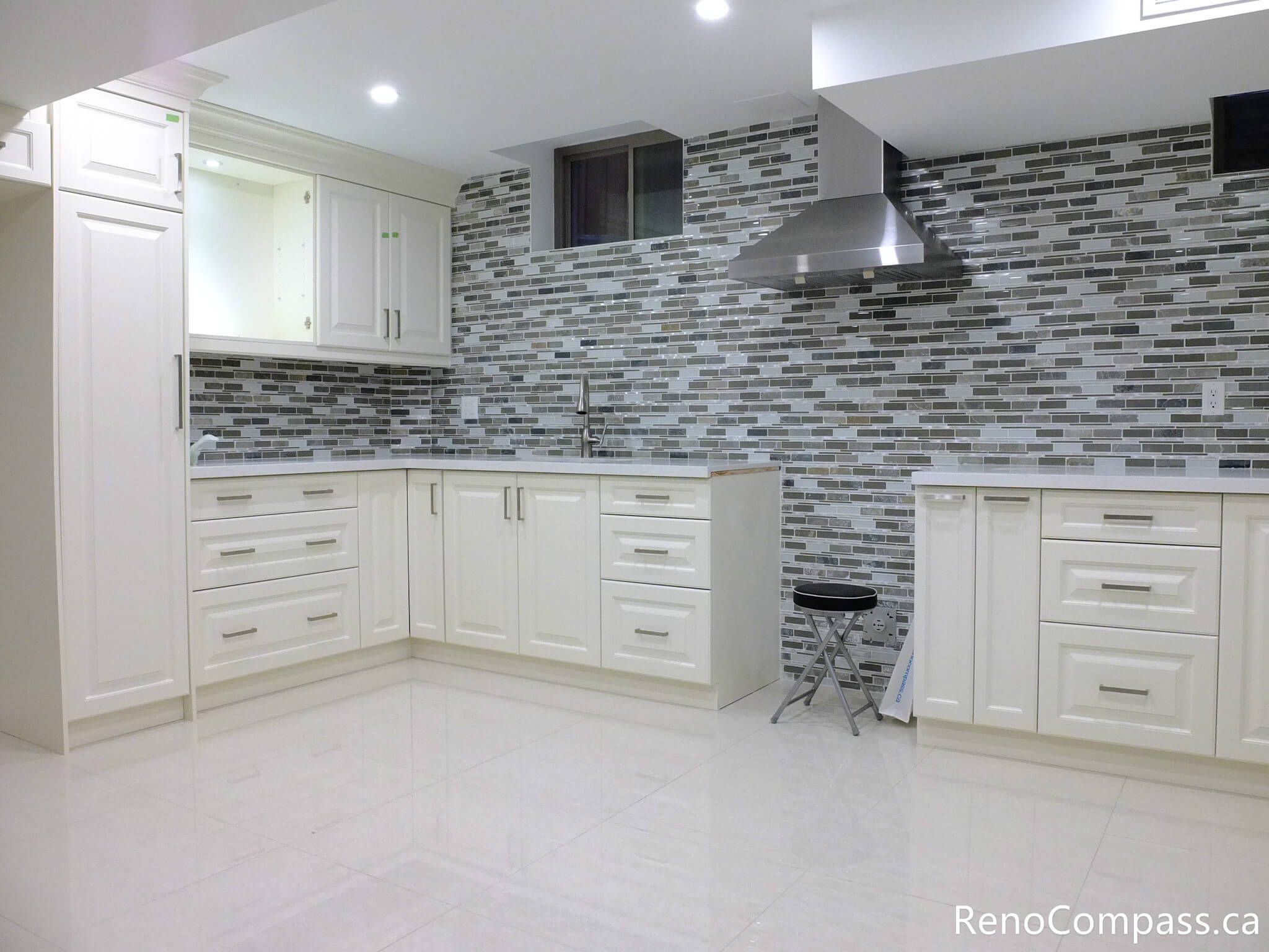 This Markham basement renovation features a custom L-shaped kitchen with classic white shaker cabinets, stainless steel range hood, and modern mosaic tile backsplash. The clean layout, bright lighting, and durable flooring create a functional and stylish secondary living space. Completed by a trusted Toronto renovation company, this project highlights quality craftsmanship and smart basement design solutions.