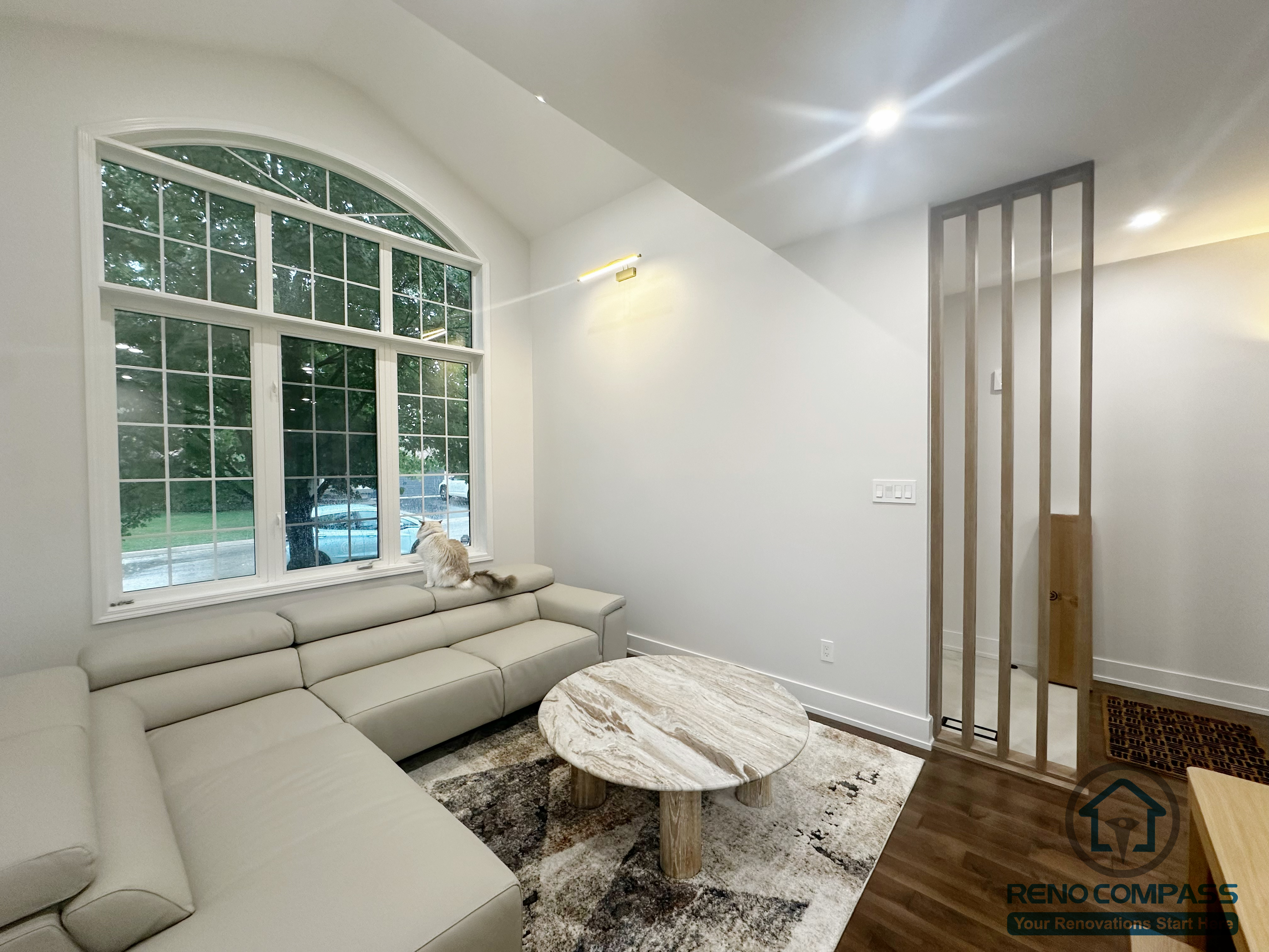 This Oakville interior renovation features a refined living room designed around natural light and comfortable daily use. A large arched window becomes the visual focal point, bringing in abundant daylight and enhancing the sense of openness. The layout pairs a neutral sectional sofa with a marble coffee table and soft area rug, creating a balanced and inviting seating area. Clean wall finishes, recessed lighting, and warm wood flooring contribute to a calm and cohesive atmosphere. The space is thoughtfully arranged to support relaxation, conversation, and long-term livability within a modern home.