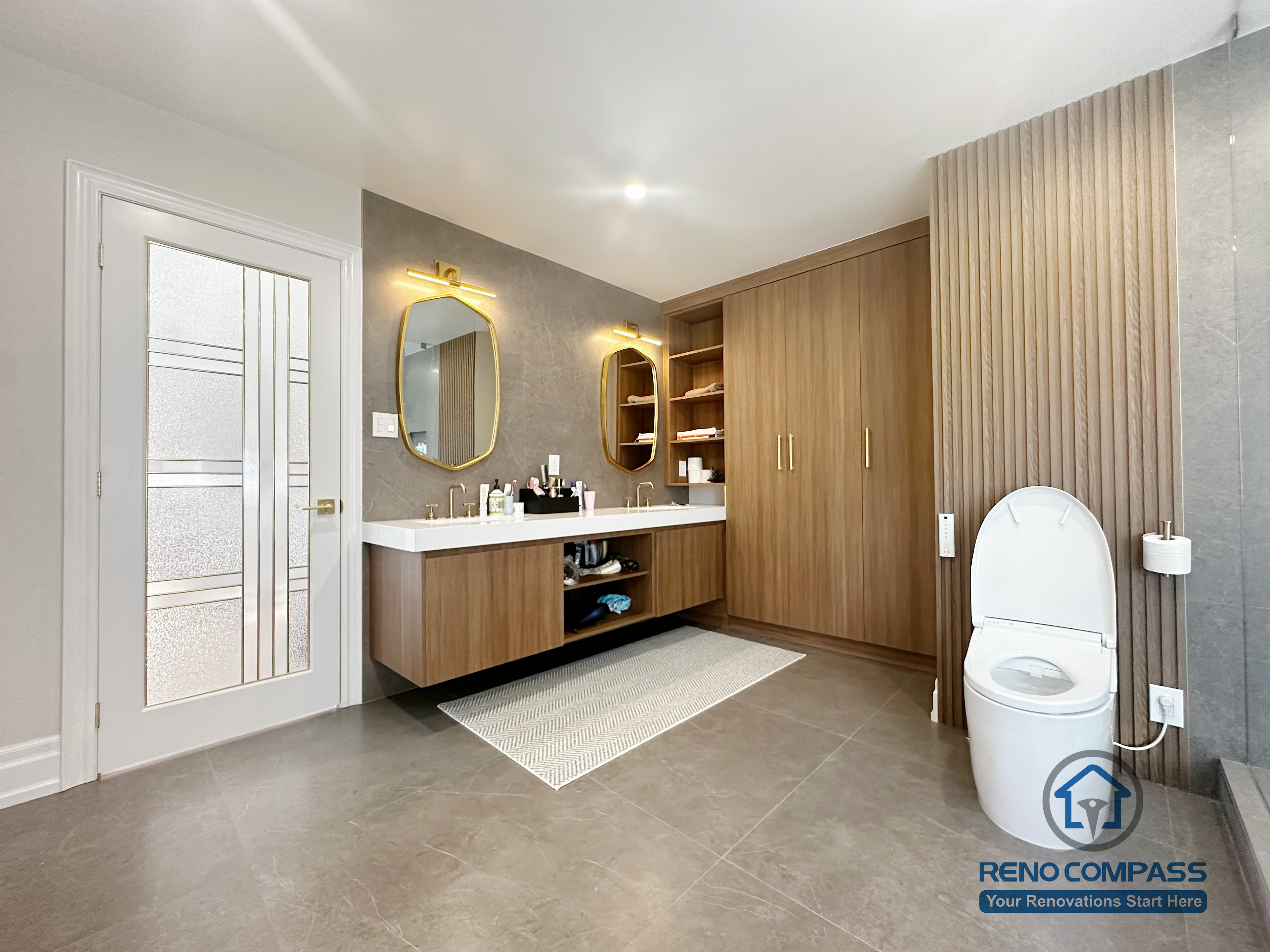 This image showcases a modern bathroom renovation completed in North York, featuring a spacious walk-in glass shower, custom wood vanity cabinetry, and built-in storage solutions. Large-format floor tiles enhance durability and visual continuity, while the frameless glass enclosure keeps the space open and bright. The combination of warm wood tones and neutral finishes creates a balanced, contemporary bathroom design focused on comfort, functionality, and everyday usability in North York homes.