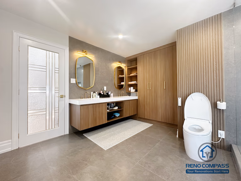 This image showcases a modern bathroom renovation completed in North York, featuring a spacious walk-in glass shower, custom wood vanity cabinetry, and built-in storage solutions. Large-format floor tiles enhance durability and visual continuity, while the frameless glass enclosure keeps the space open and bright. The combination of warm wood tones and neutral finishes creates a balanced, contemporary bathroom design focused on comfort, functionality, and everyday usability in North York homes.