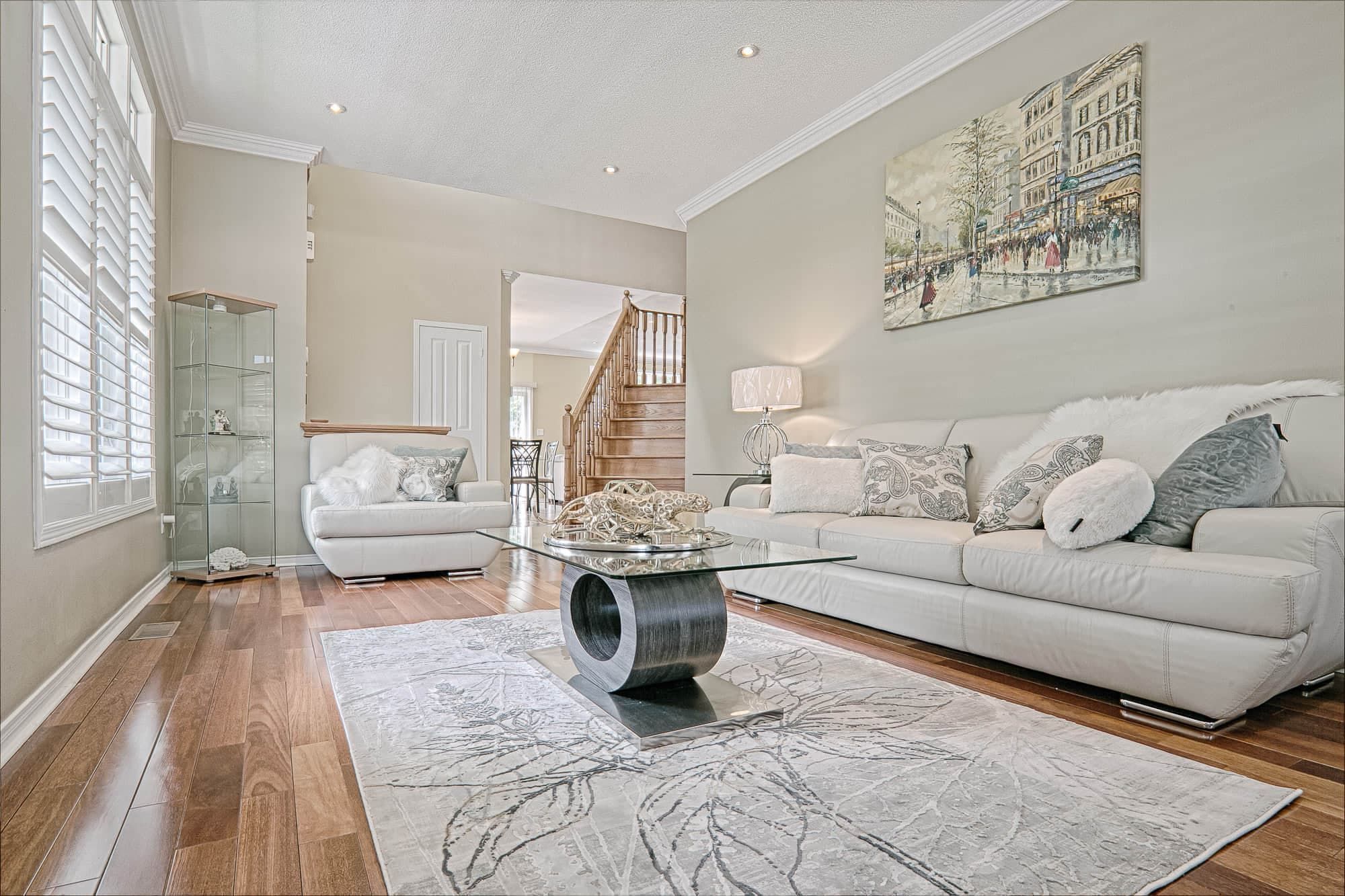 This elegant living room interior showcases warm hardwood flooring, soft neutral wall tones, and coordinated modern furniture that create a bright and sophisticated atmosphere. The large bay windows with plantation shutters allow abundant natural light, while the contemporary coffee table, textured area rug, and layered lighting enhance comfort and style. A refined example of warm minimalist living room renovation and modern home interior design inspiration for homeowners in Toronto and the GTA.