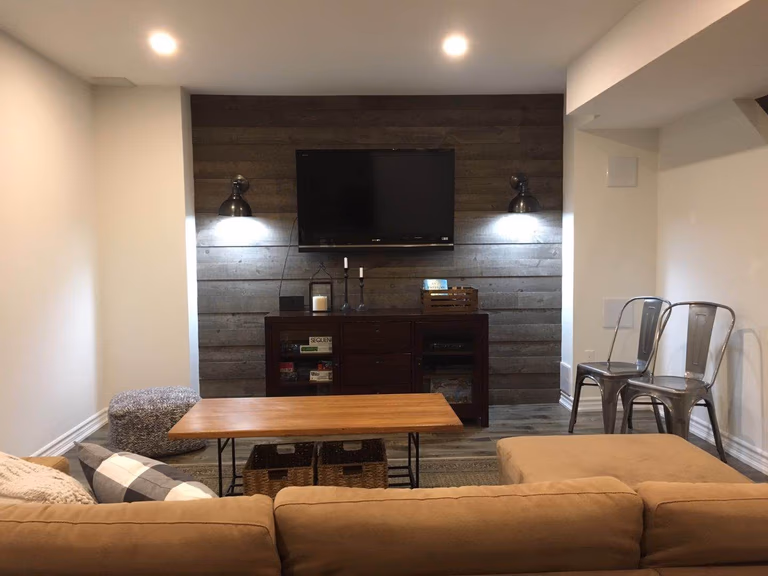 This Richmond Hill basement renovation highlights a beautifully designed entertainment space featuring a custom TV feature wall with wood panel accents, modern wall sconces, and built-in storage. The finished basement living area offers a warm and inviting atmosphere with contemporary furnishings, upgraded flooring, and functional layout, creating the perfect space for family relaxation and entertainment.