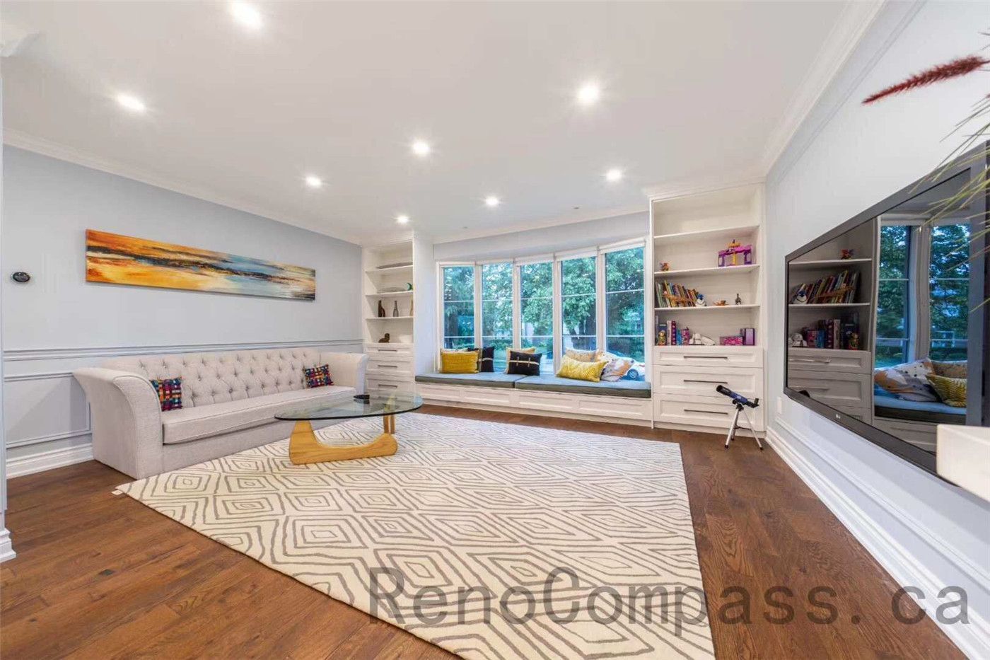 This modern living room is part of a North York interior renovation, designed to balance comfort, functionality, and visual openness. Custom built-in shelving frames the seating area, while recessed ceiling lights provide even illumination throughout the space. A large picture window with a built-in bench brings in natural light and creates a cozy reading corner. Warm hardwood flooring, neutral finishes, and a clean layout reflect the quality craftsmanship and thoughtful space planning typical of professional Toronto interior renovation projects.