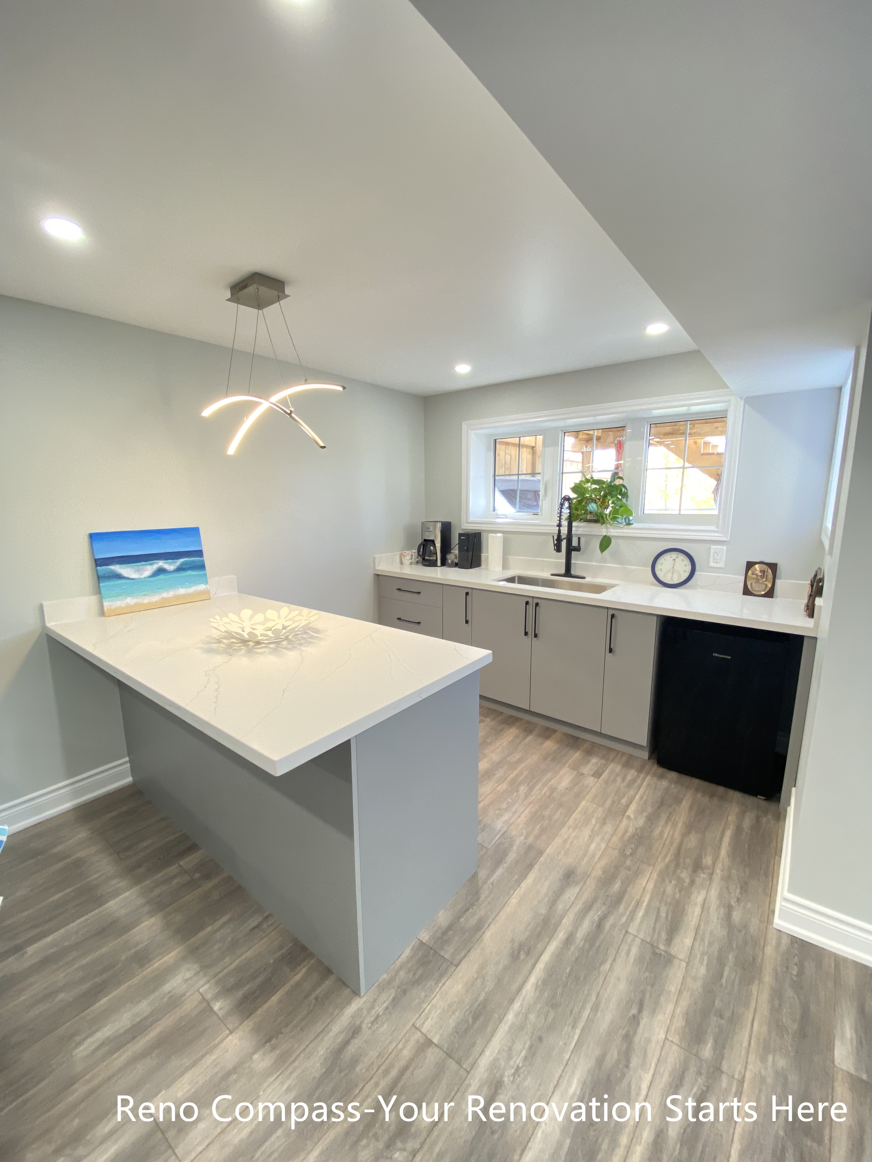 Modern basement bar renovation in Oakville featuring a large quartz island countertop, custom base cabinets, contemporary pendant lighting, vinyl plank flooring, and a compact kitchenette layout designed for entertaining and everyday use, completed by Reno Compass Inc.