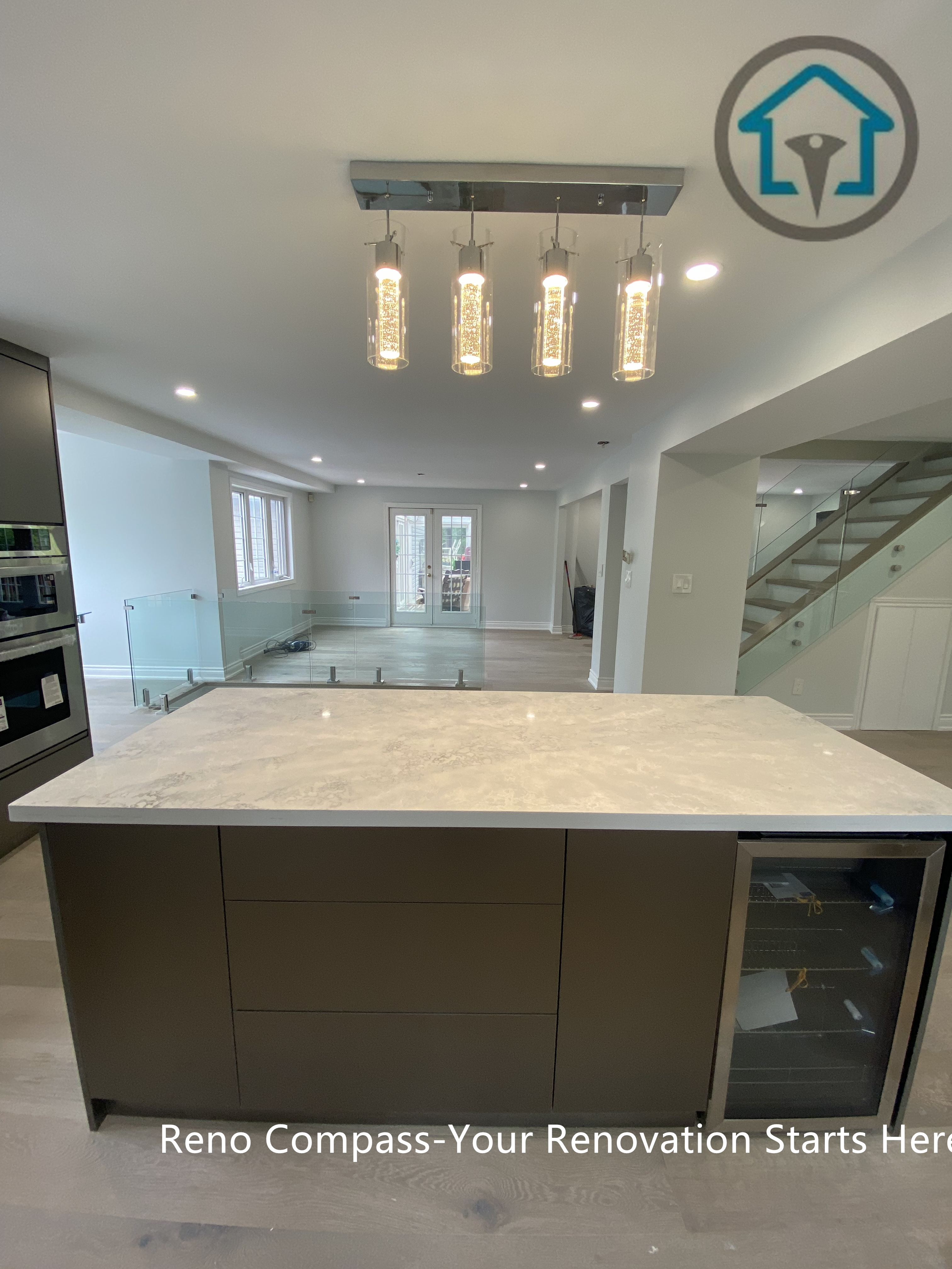 This view highlights the custom kitchen island featuring a seamless stone countertop, built-in storage, and integrated wine fridge. Linear pendant lighting and recessed ceiling lights provide balanced illumination, while the open sightlines connect the kitchen to the main living area and staircase, reinforcing the home’s modern open-concept design.