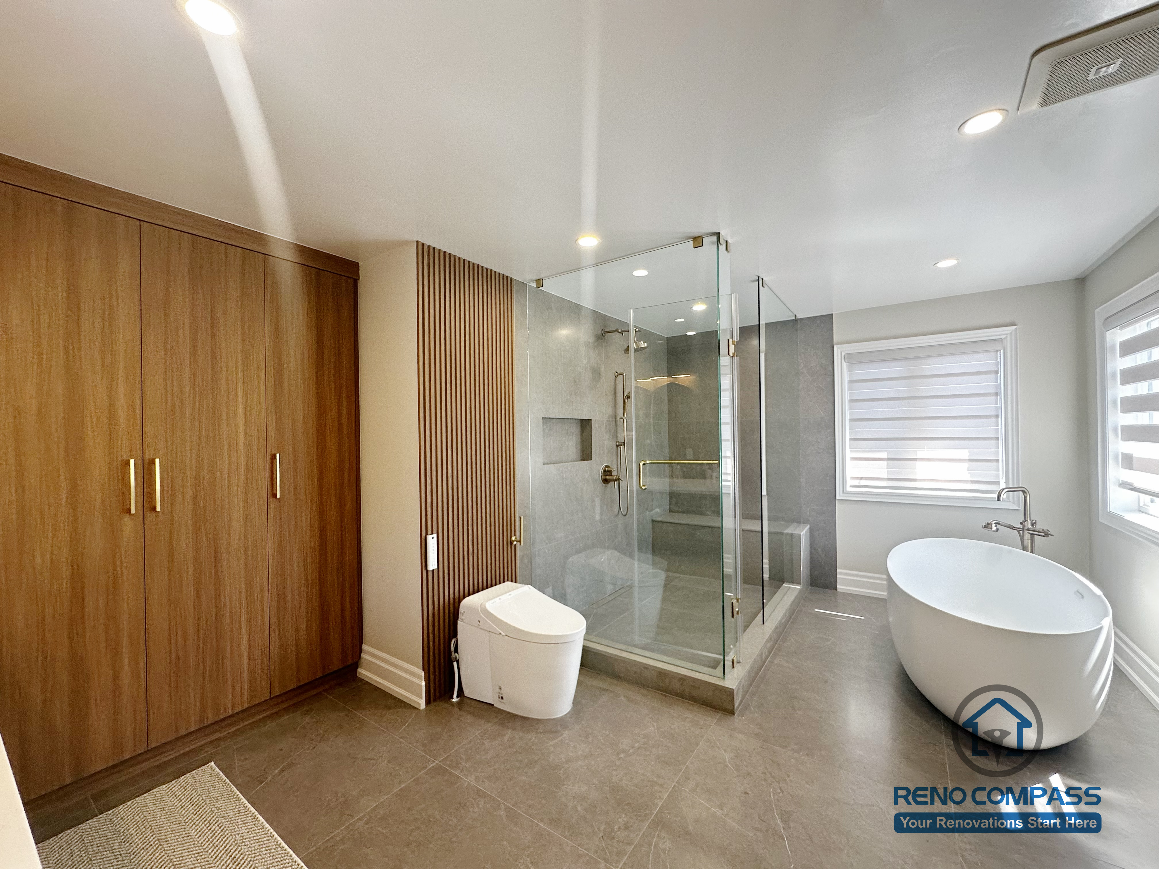 This image showcases a spacious primary bathroom renovation completed in North York, featuring a walk-in glass shower with built-in niche, a freestanding soaking tub, and custom wood storage cabinetry. Large-format floor tiles provide durability and visual continuity, while the frameless shower enclosure enhances openness and natural light. The layout balances functionality and comfort, creating a modern bathroom designed for everyday use and long-term living in North York homes.