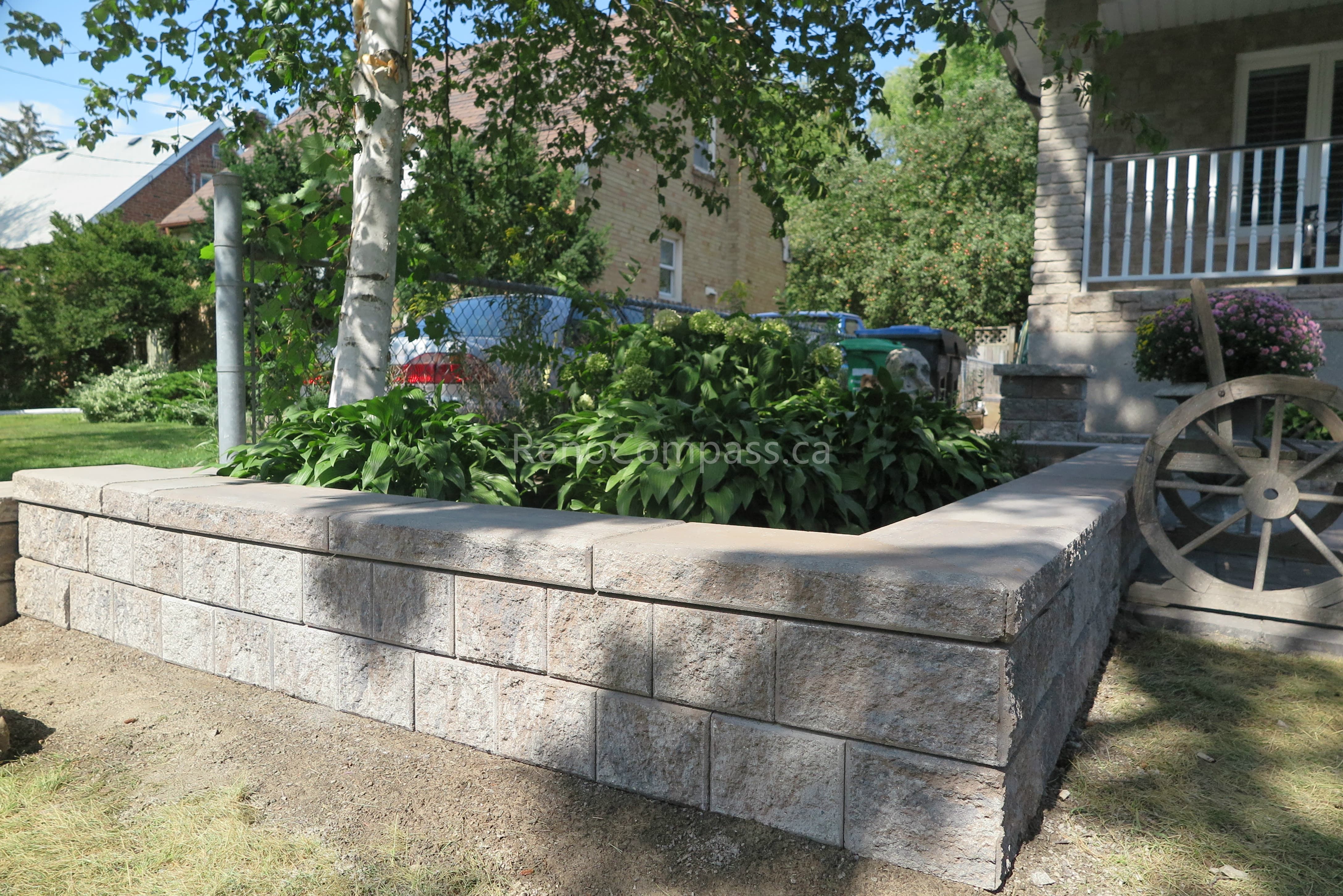 This close-up view highlights the precision craftsmanship of the stone retaining wall and interlocking patio installation in Mississauga. The textured concrete blocks and smooth stone cap provide structural stability while delivering a clean, layered aesthetic. Integrated seamlessly with the surrounding landscaping and planting design, this hardscape feature enhances both durability and visual depth. Professionally completed by Reno Compass Inc., this project reflects expert workmanship in outdoor interlocking and landscape construction.